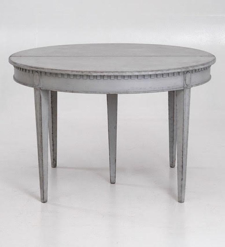 A beautiful Swedish three-leaf oval dining table in the Gustavian Style with a light gray patina. There is beautifully carved dentil molding around the apron with a carved oval motif above each square, tapered, and fluted leg. This is a charming and