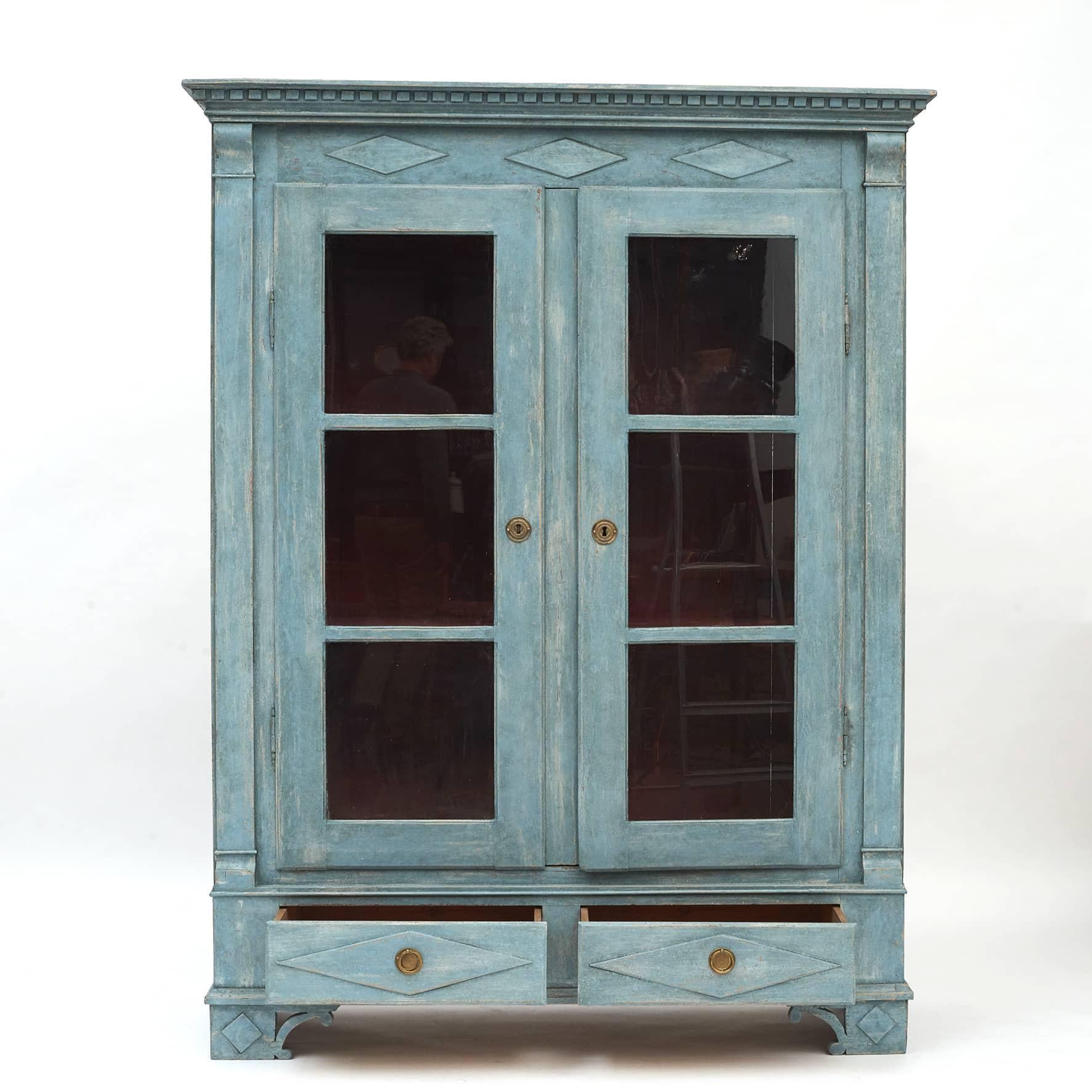 Swedish Gustavian style vitrine/display cabinet.
Pine wood later professional painted blue with wonderful distressed finish, the interior painted in a dark Pompeii red.
Cornice featuring carved dentil and rhombuses molding. Pair of glass doors