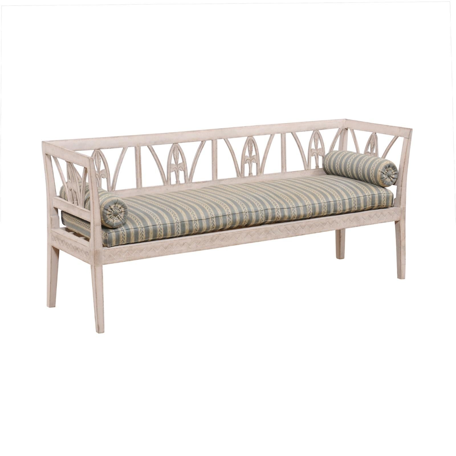 A Swedish Late Gustavian period painted wood sofa bench from the early 19th century with foliage-carved back, jagged motifs and upholstery. Created in Sweden during the first quarter of the 19th century, this Late Gustavian bench features a