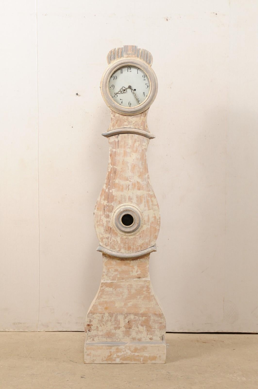A Swedish grandfather clock with scraped finish from the 19th century. This antique clock from Sweden features a low profile reed carved crown adorning it's head, a teardrop-shaped belly with window at it's center, and is supported upon a flat