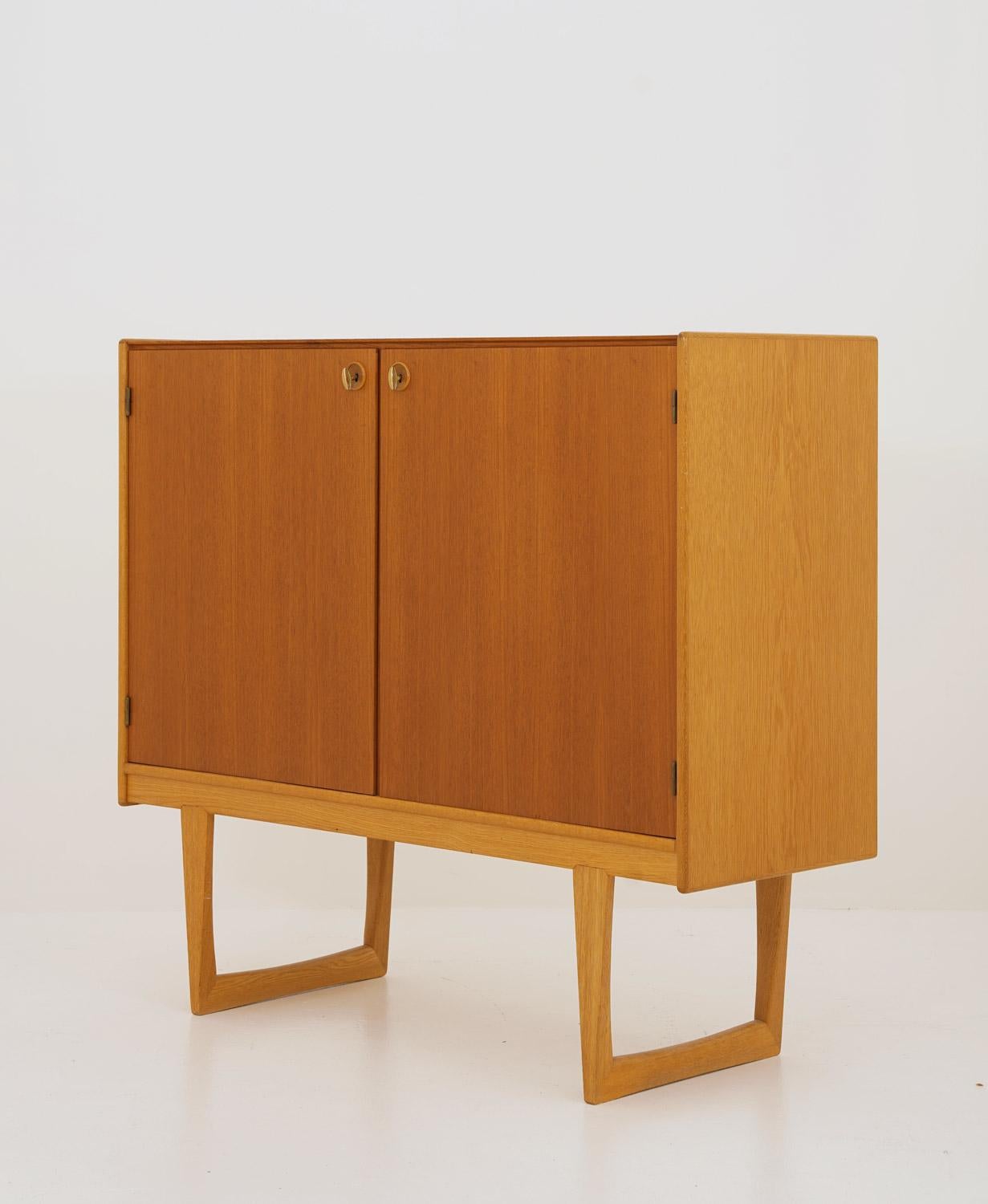 Elegant and minimalistic cabinet by Yngvar Sandström for Nordiska Kompaniet (NK). The cabinet features two doors in teak with original brass keys. The legs and the sides are made of oak, creating a beautiful contrast between the different types of