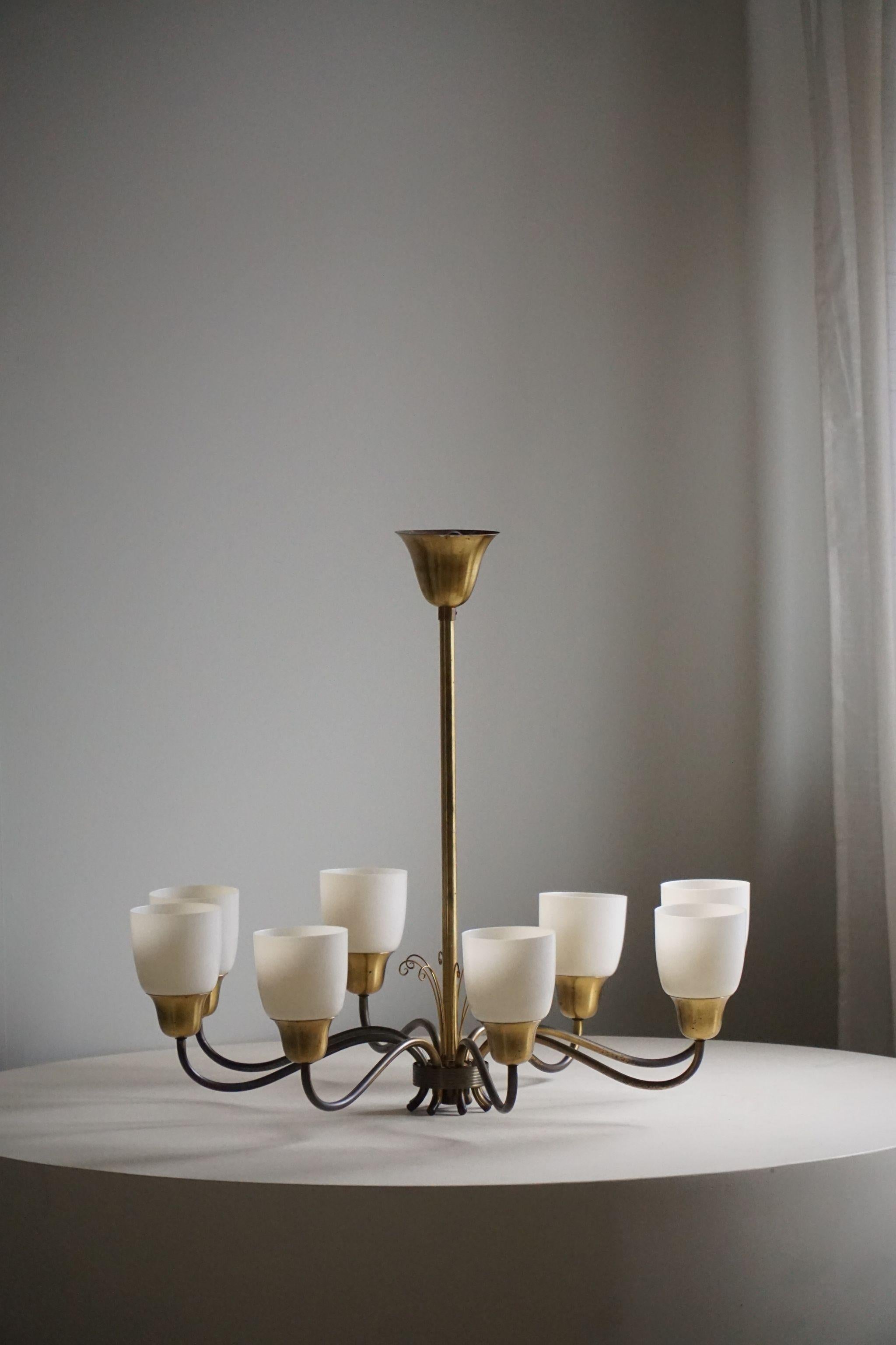 This Swedish Mid Century Modern chandelier from the 1950s features eight gracefully curved brass arms, each adorned with a plastic shade. With its minimalist design and clean lines, it embodies the sleek elegance characteristic of the era. Crafted