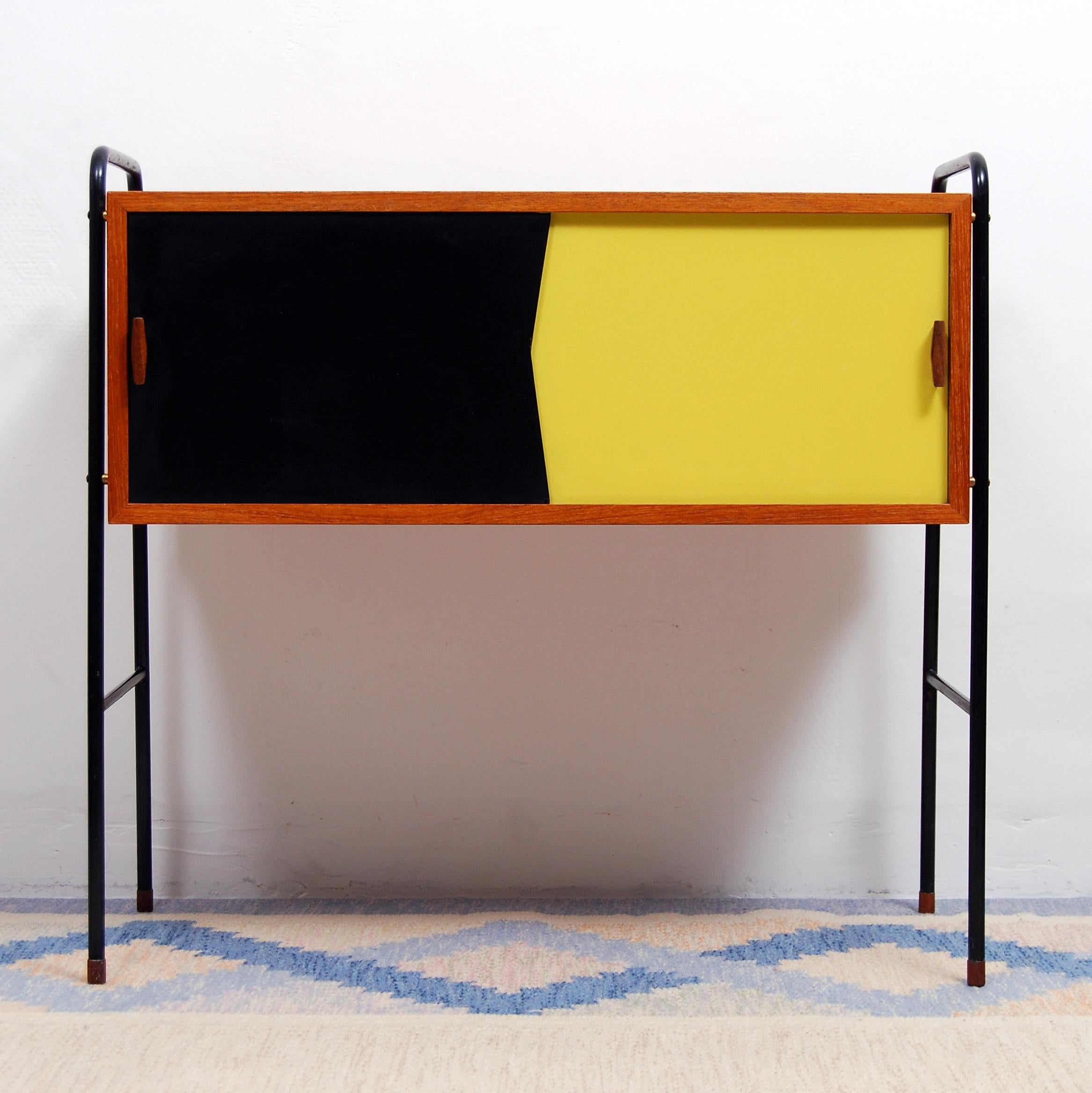 Small 1950s sideboard / cabinet in teak. Black lacquered metal legs. Black and yellow lacquered sliding front doors. Two internal sliding shelves.
  