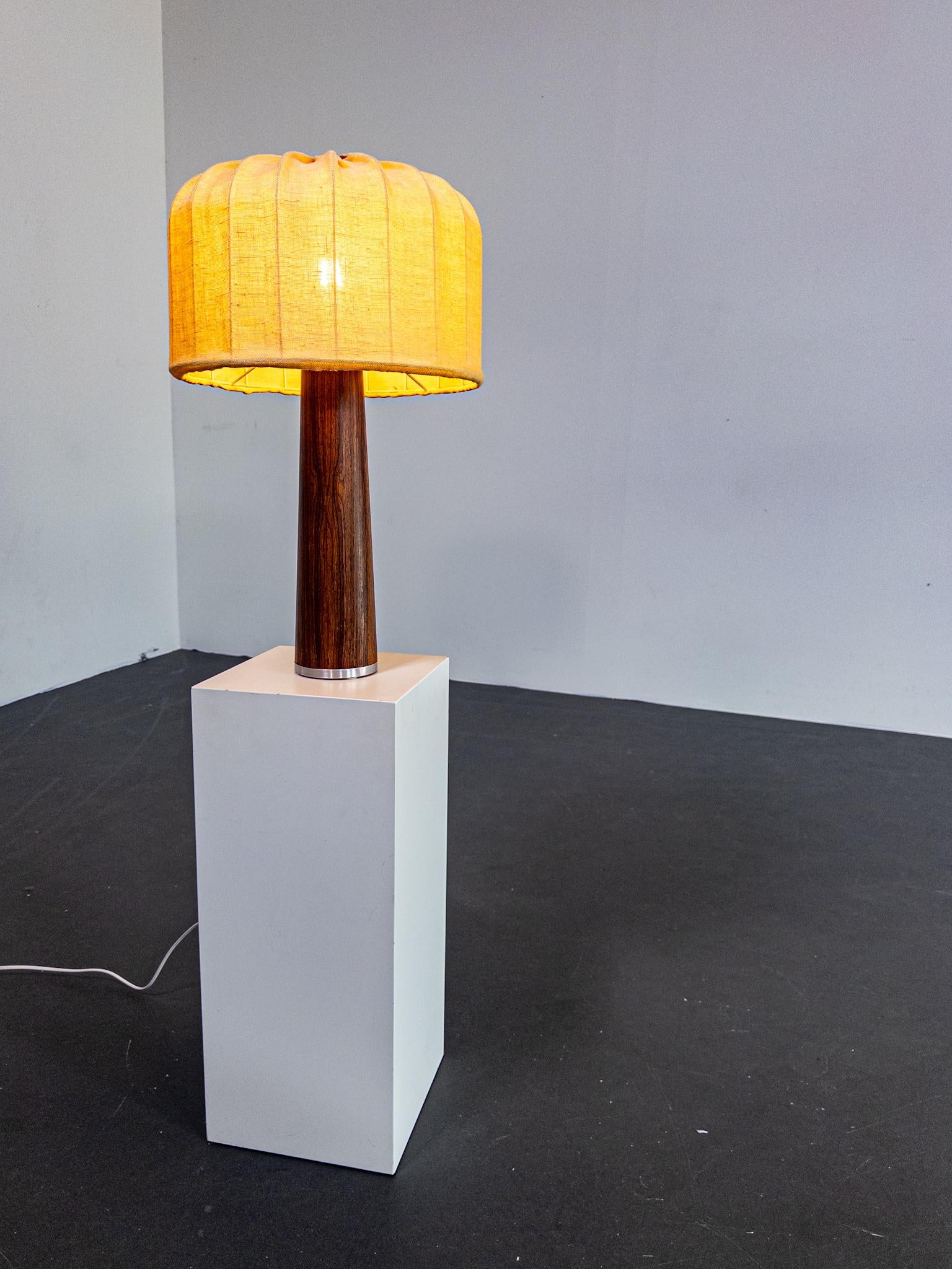 Elegante lampada da tavolo scandinava degli anni '60, realizzata in legno di palissandro riccamente venato con una raffinata silhouette conica. Il design è accentuato da un anello minimalista in acciaio inossidabile alla base, un dettaglio che