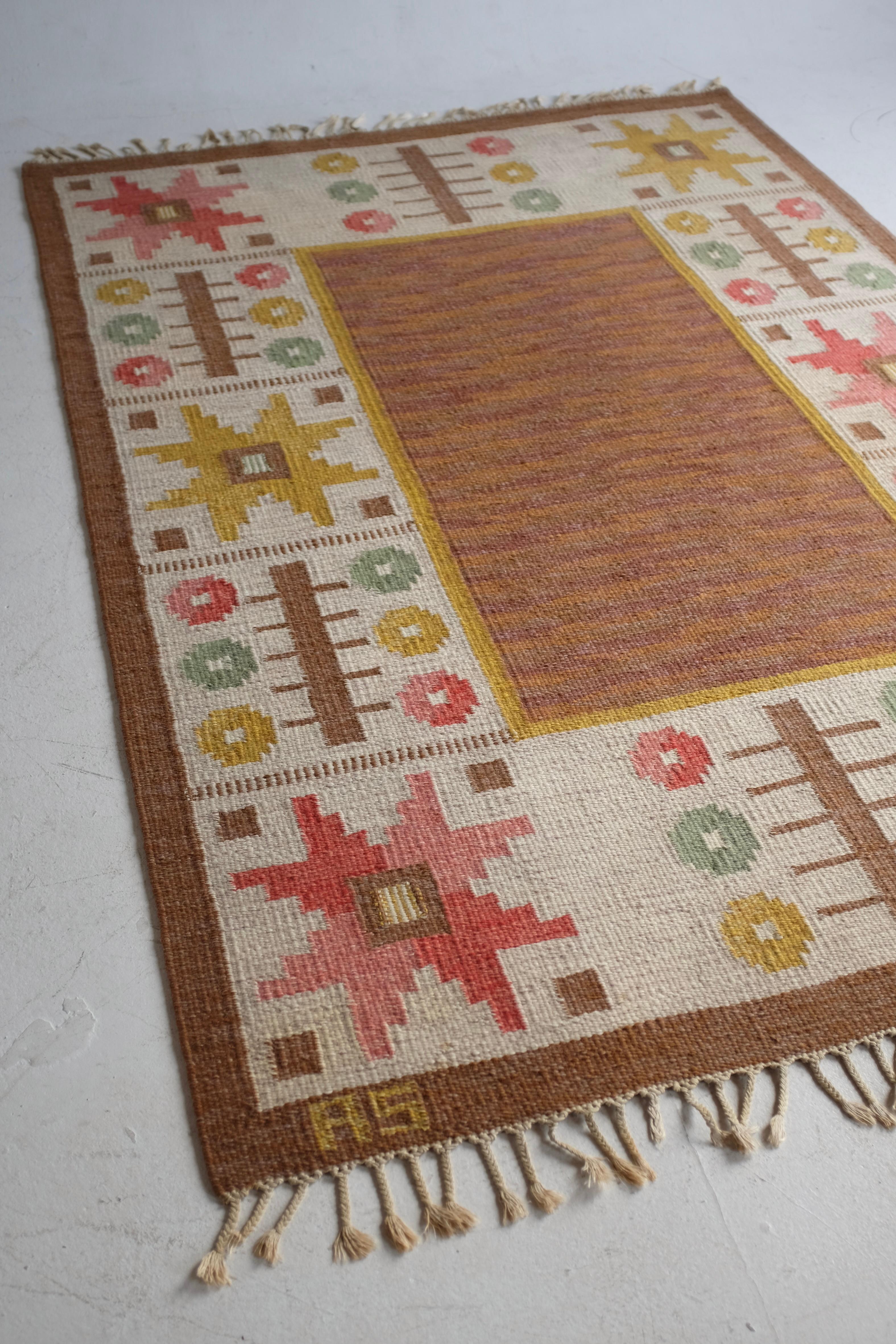 Beautiful Mid-Century rug by Swedish designer Astrid Sampe. A brown and yellow rectangular shape in the middle is surrounded of stars and organic shapes in green, yellow and pink tones. Astrid Sampe was a prolific textile designer not only in Sweden