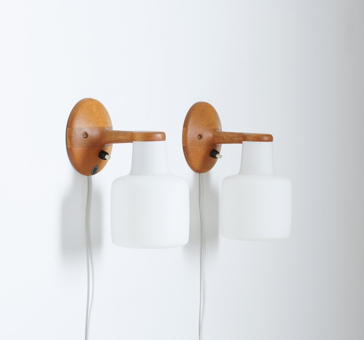 Wall lamps by Uno & Östen Kristiansson for Luxus, Sweden.
The lamps are made of oak with white opaline glass shades and give a soft, ambient light when lit. 

Condition: One lamp has a slight crack in the wood, hardly visible, that has been