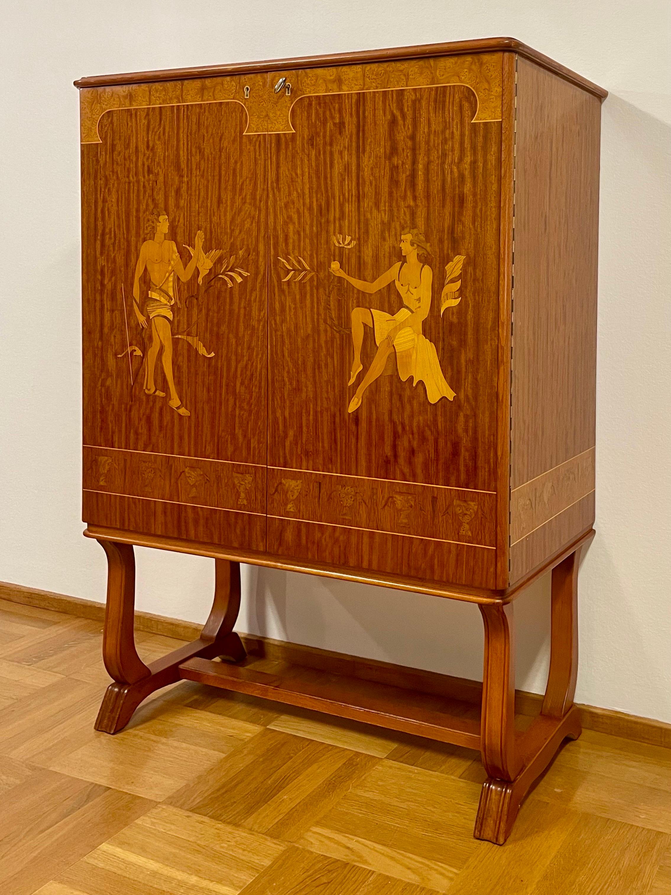 This is the Swedish Modern 1930s Cabinet with Mjölby Intarsia-decor from Vingåkers Möbelfabrik. 
It comes in very good condition, made of birch. Veneered with mahogany and decorated with inlays from Mjölby Intarsia.
Legs in stained birch. 

Behind