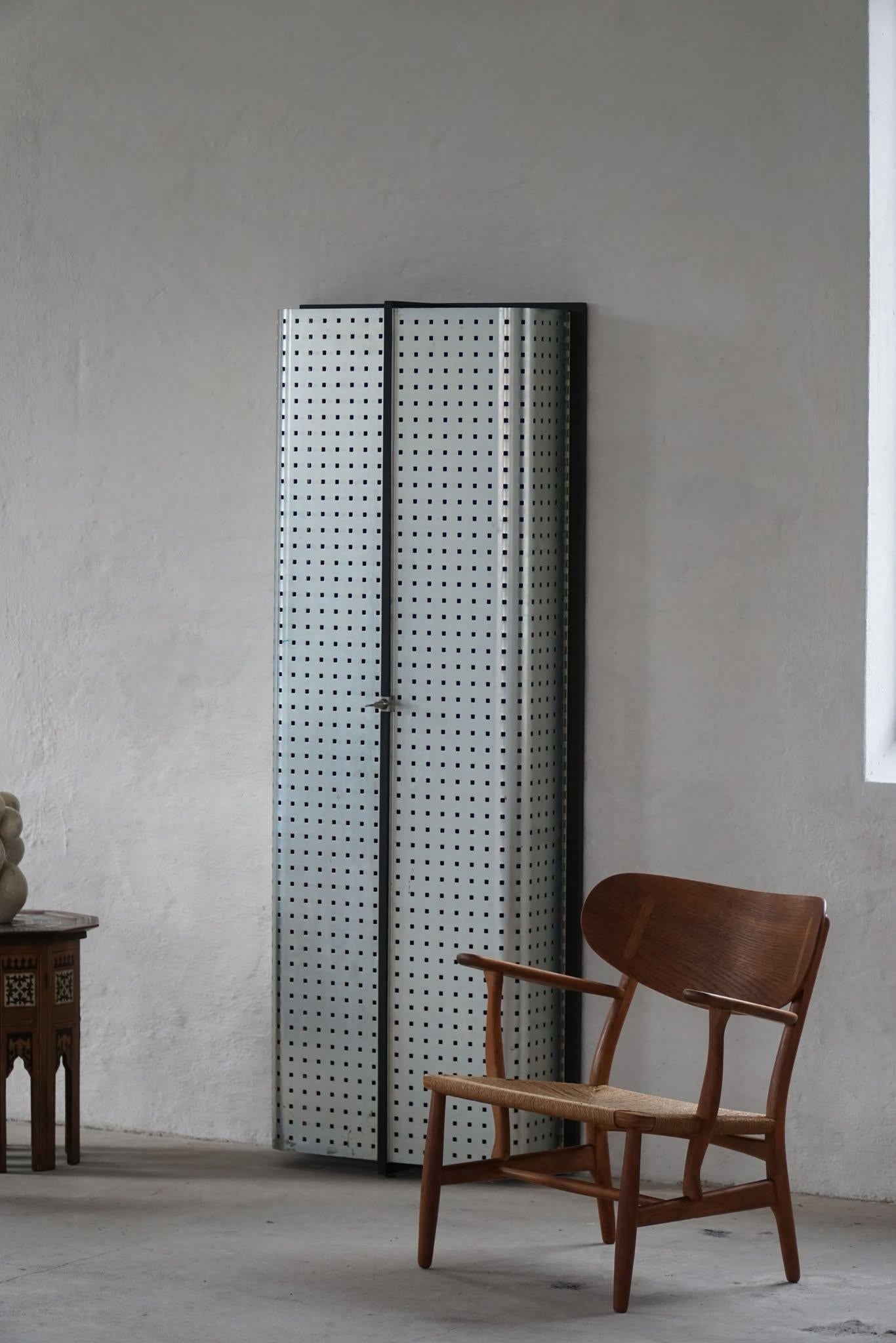 A tall, narrow Brutalist cabinet crafted from steel, featuring clean lines and an industrial aesthetic. Its minimalist frame is complemented by glass shelves, creating a striking contrast between solid metal and transparent surfaces. Made in the