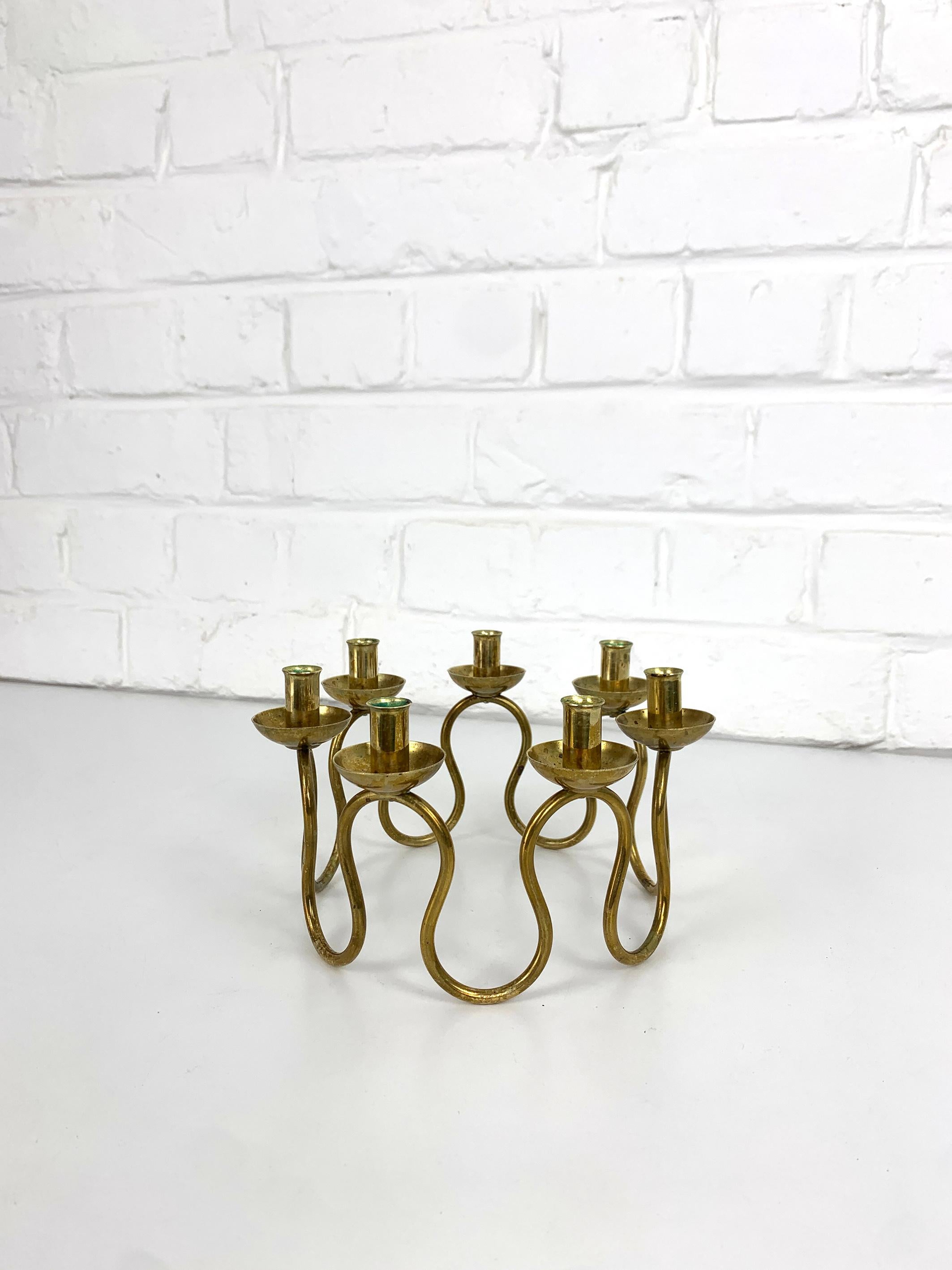A striking candelabra in solid brass, designed by Lars Holmström for Svenskt Tenn. This sculptural piece features a sinuous, tubular structure rising from a circular base, supporting seven cylindrical candle holders in a harmonious rhythm. The