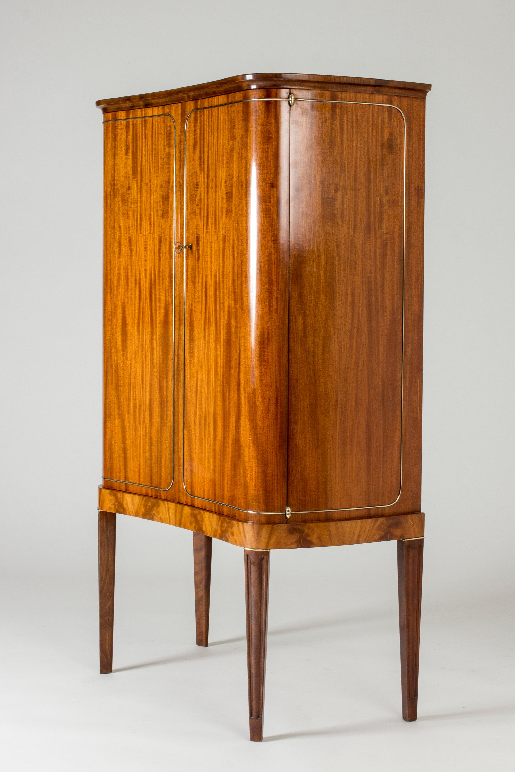 Stunning Swedish Modern cabinet by Axel Bäck. Made from mahogany. Beautifully arched front with inlayed decor of a brass line, tapering legs. Carefully executed through and through, with neat metal handles on the drawers on the inside.