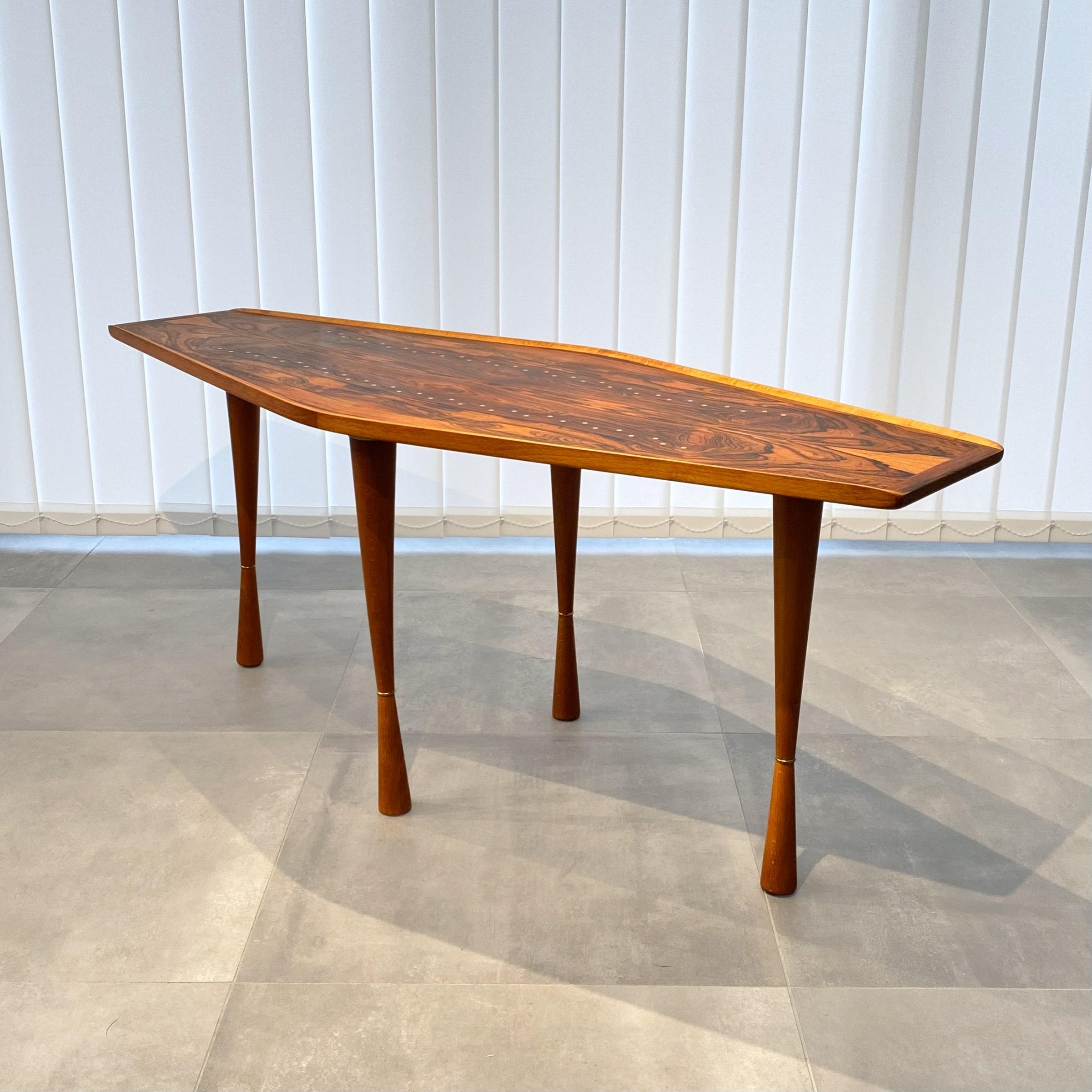 Swedish 1940s modernist coffee table constructed from a hexagonal tabletop with upturned edges resting on four hourglass-shaped legs with brass ring decorations. The tabletop is adorned with brass inlays, reinforcing the dramatic appearance of the