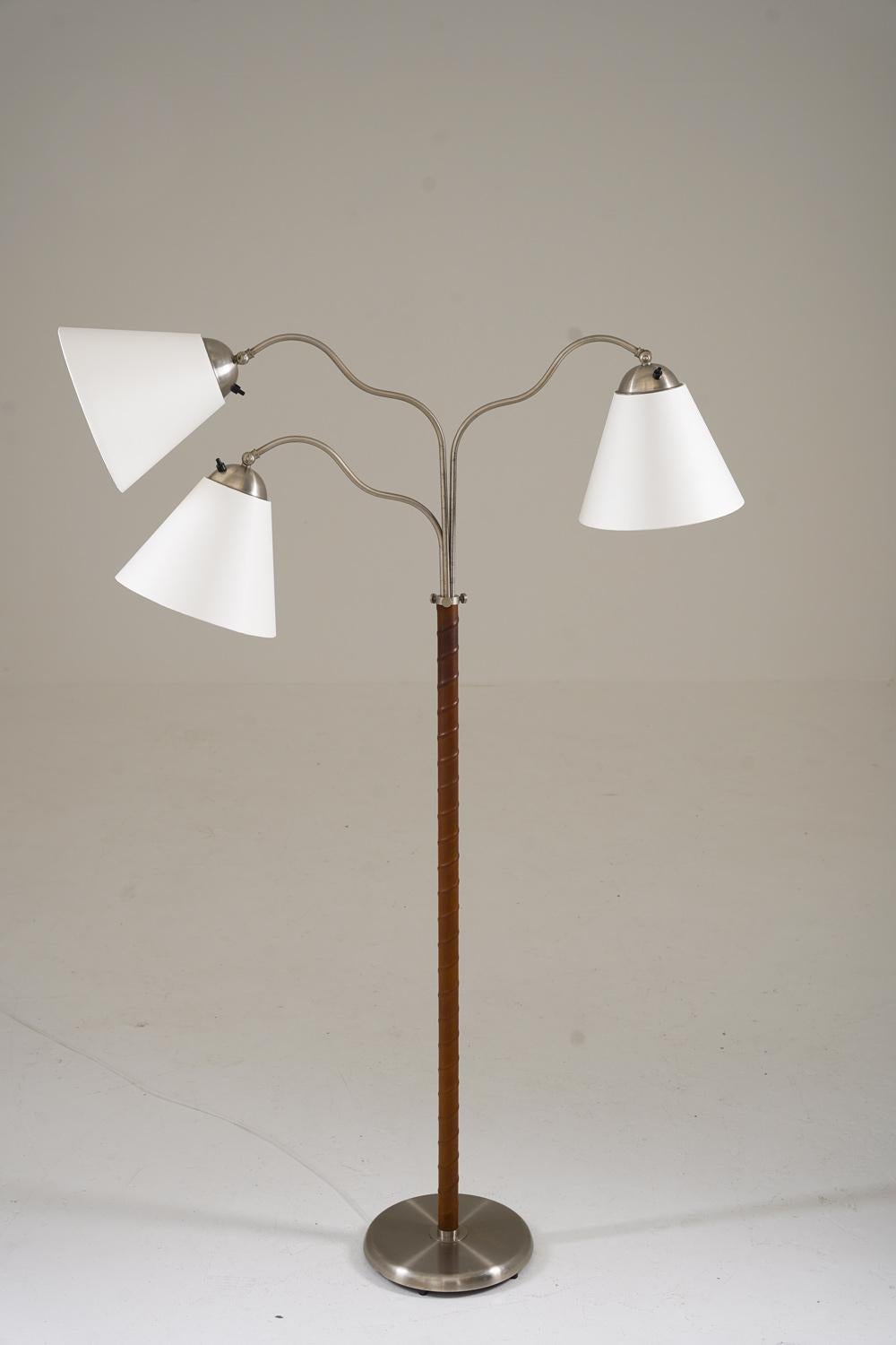 This is a lovely Swedish Modern floor lamp that was manufactured in Sweden during the 1940s. The lamp features a brushed metal base and a rod with leather webbing. The rod supports three swivel arms that hold the shades. The swivel arms are
