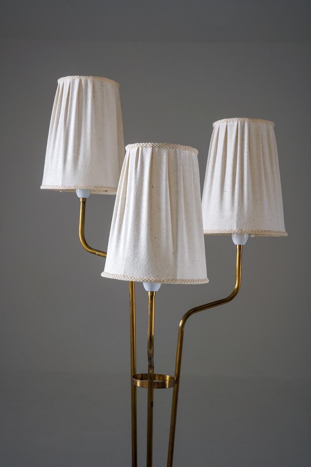 A graceful Swedish floor lamp from the 1940s, featuring a brass construction with three elegantly curved arms rising from a circular base. Each arm supports its original pleated fabric shade, creating a balanced composition and a warm, diffused