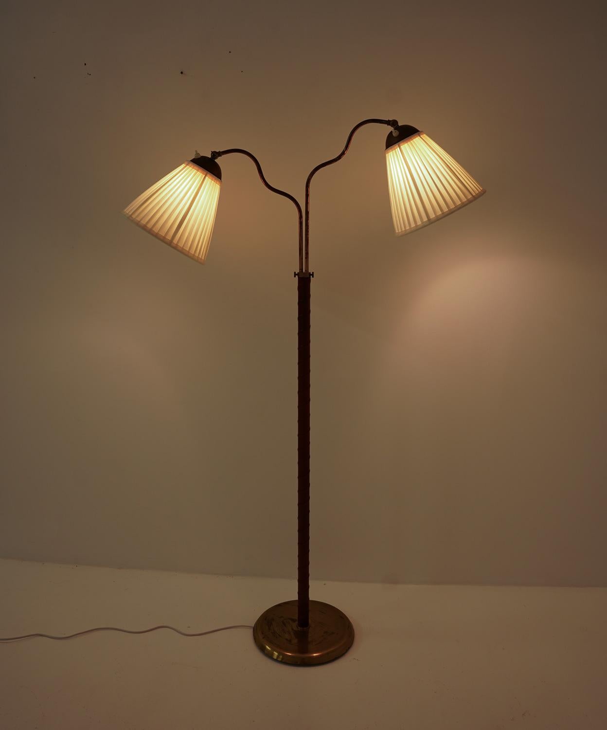 This is a lovely Swedish Modern floor lamp that was manufactured in Sweden during the 1940s. The lamp features a brass covered iron base and a brass rod with leather webbing. The rod supports two swivel arms that hold the shades. The swivel arms are