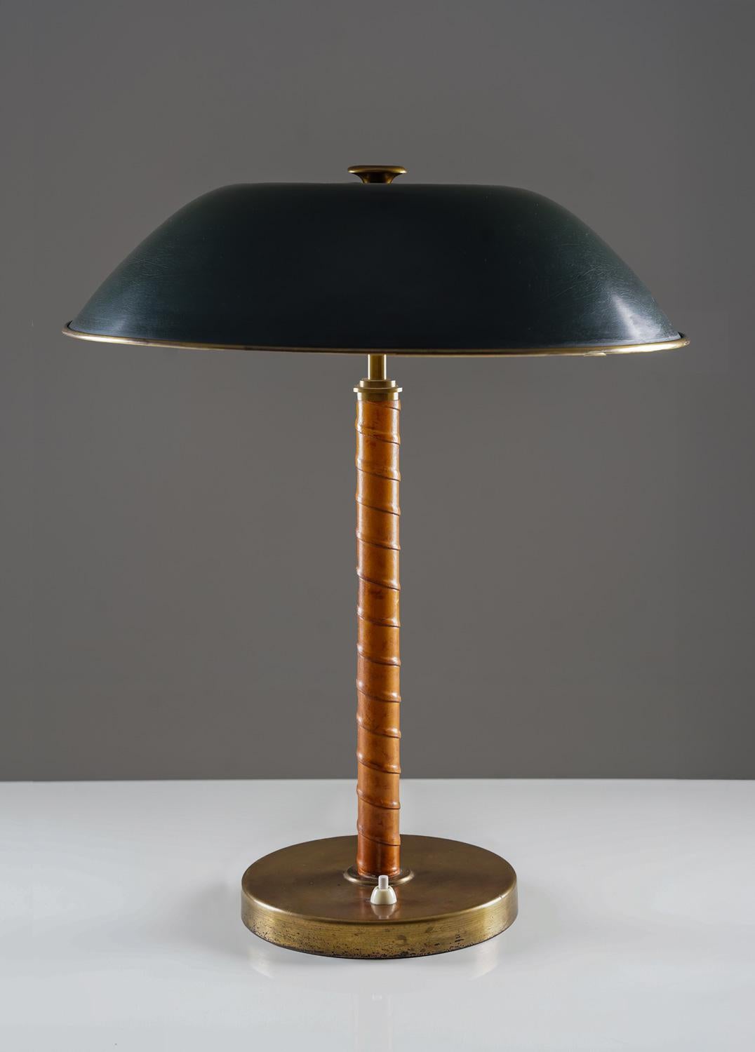 A striking table lamp by Böhlmarks, model 15624, produced during the 1940s–50s. The lamp features a wide, dark green lacquered metal shade with brass details, balanced on a cylindrical stem wrapped in spiral-stitched leather, and set on a circular