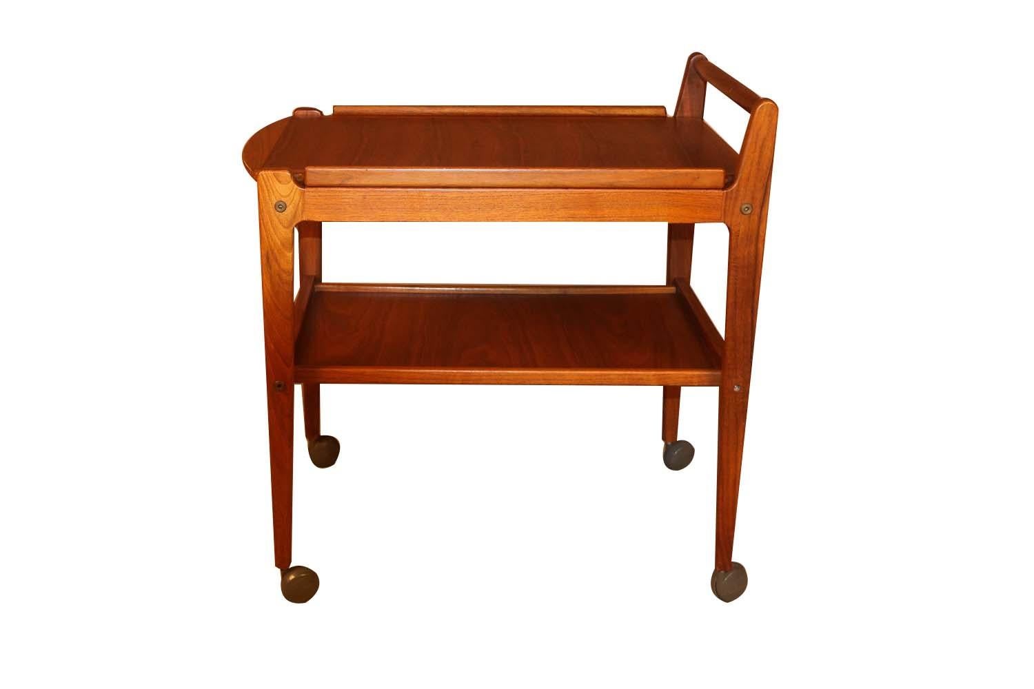 A Classic midcentury bar cart attributed to Erik Gustafsson, crafted in teak solids and veneers. The simple and functional cart often referred to as a “drink trolley”, features a removable molded tray top angled arm and legs over a fixed lower