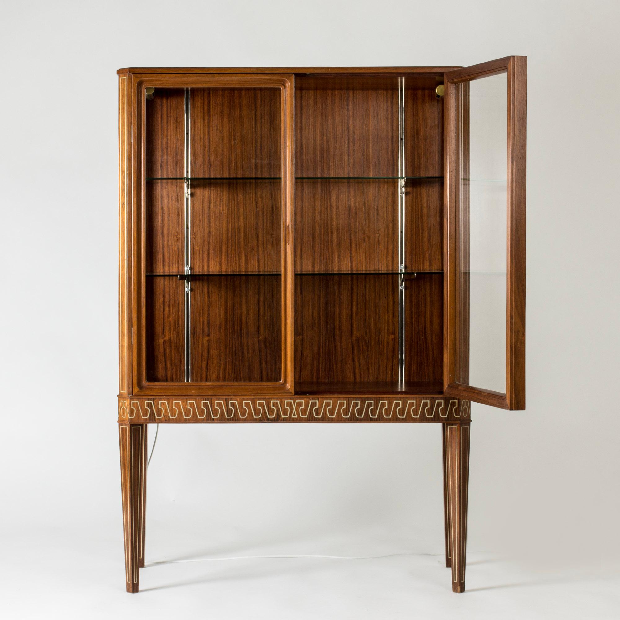 Beautiful Swedish Modern vitrine cabinet, made from rosewood with glass fronts. Elegant, graphic inlayed band along the bottom edge, made in brass. Brass decor on the legs and corners. Perfect for a cherished collection of beautiful things.