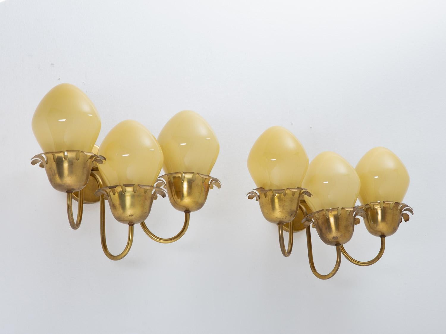 Rare wall sconces by unknown manufacturer, most likely produced in Sweden, circa 1940.
These lamps feature three light sources, hidden by a large yellow-white glass shade. The shade is held by a polished brass frame with beautiful details, typical