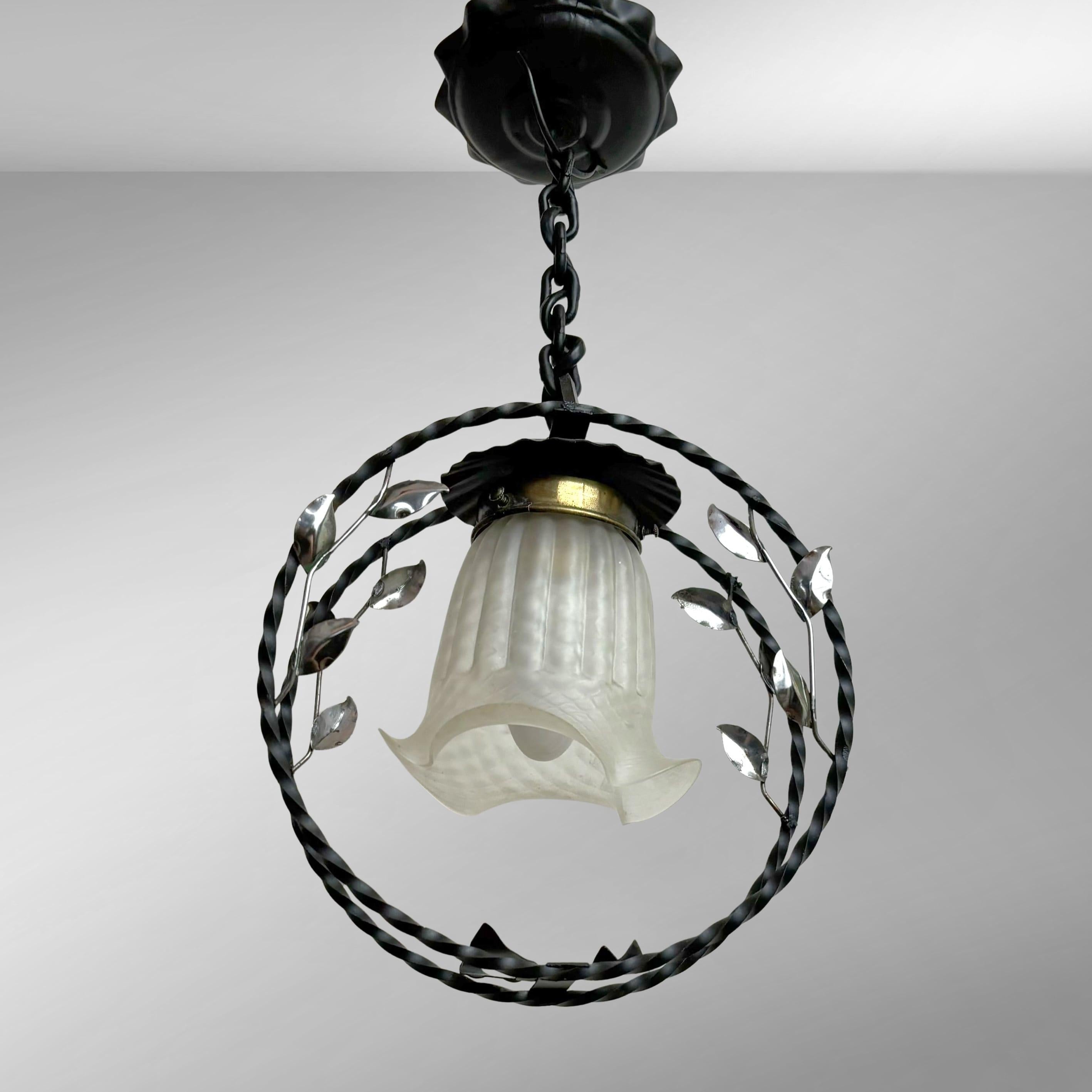 A Swedish Modern pendant lamp crafted from wrought iron, featuring a circular frame suspended by a metal chain from a matching ceiling canopy. The fixture is decorated with metal branches and leaves, with an organically shaped glass shade in the