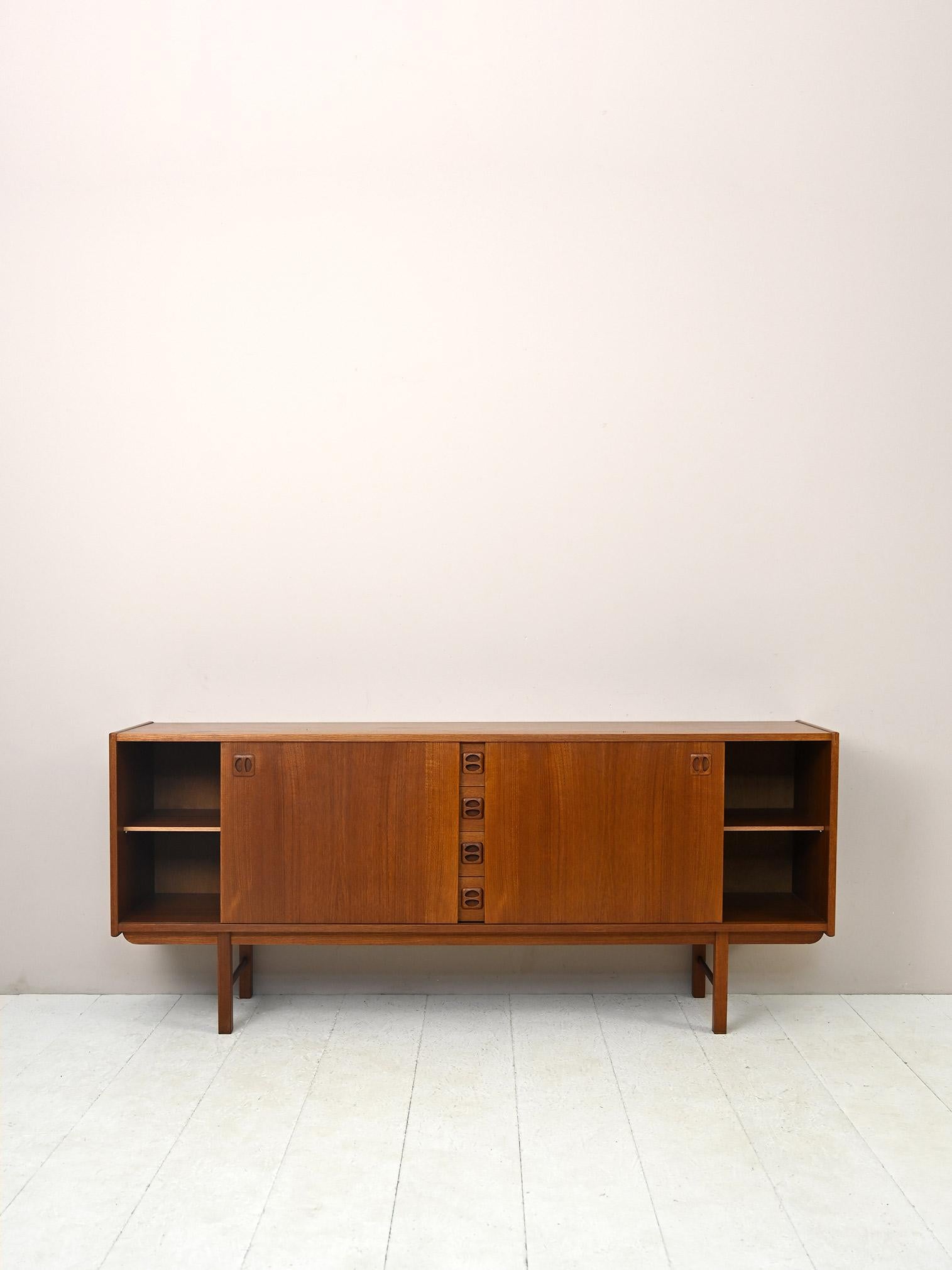Modernist sideboard of Scandinavian manufacture.

A piece of furniture with Nordic flavor and old-world warmth. The minimal and classic lines are enhanced by the carved wooden handle of the drawers and sliding doors.
The bold shade of teak wood