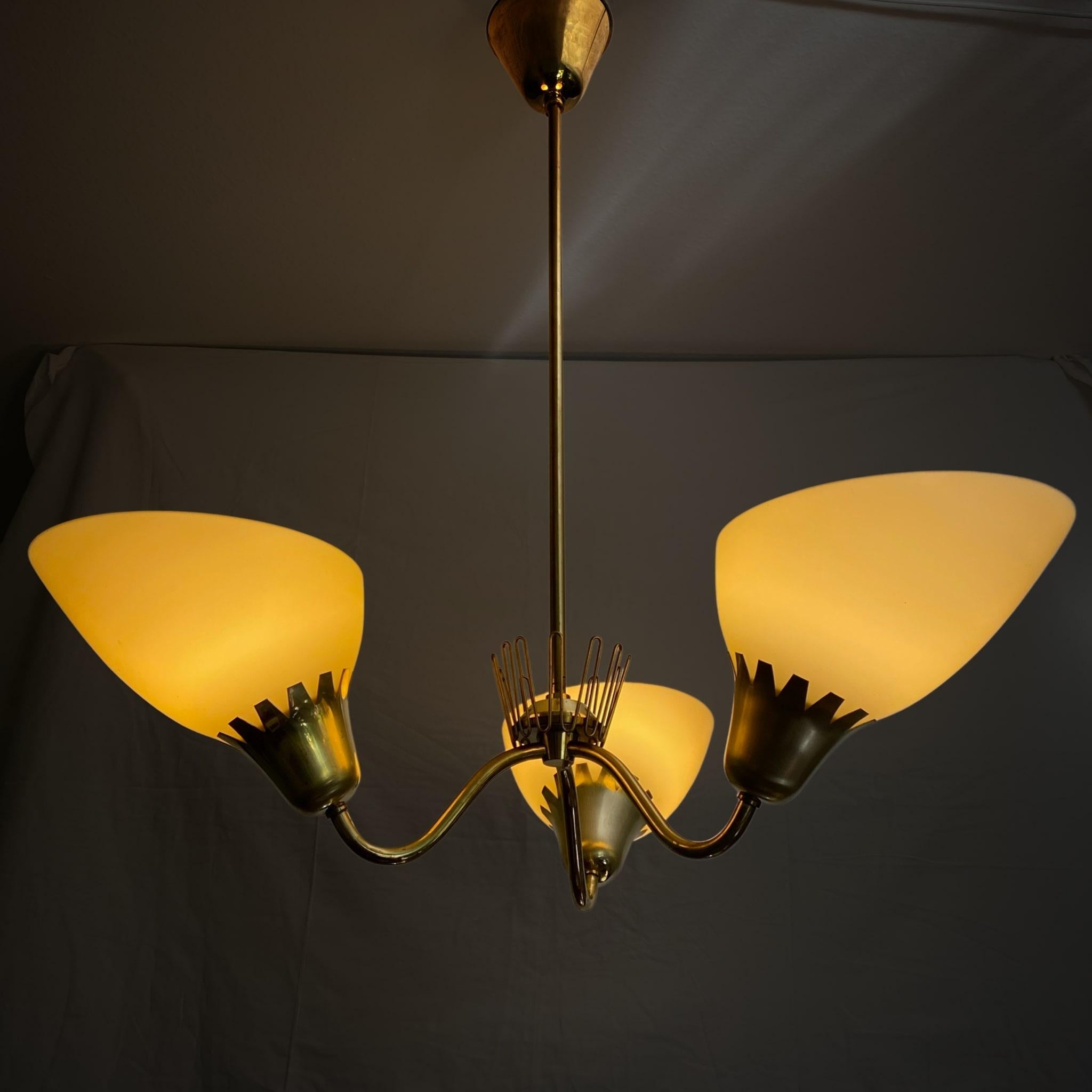 Swedish mid century chandelier model A4350 produced by the Swedish electric company ASEA in the 1950s. Sculptural frame made from brass with three arms holding amber colored glass shades. Decorative wave shaped ornament in the center of the fixture