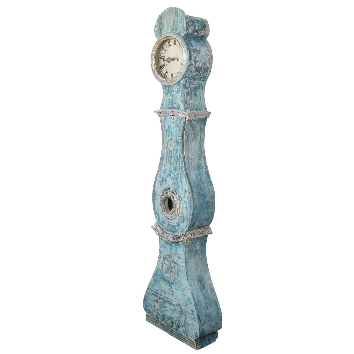 Un reloj sueco de caja larga bellamente envejecido en el clásico estilo Mora, con una silueta graciosamente curvada y un acabado pintado de azul desgastado. La esfera con números romanos y los detalles tallados a mano captan el encanto rústico y la