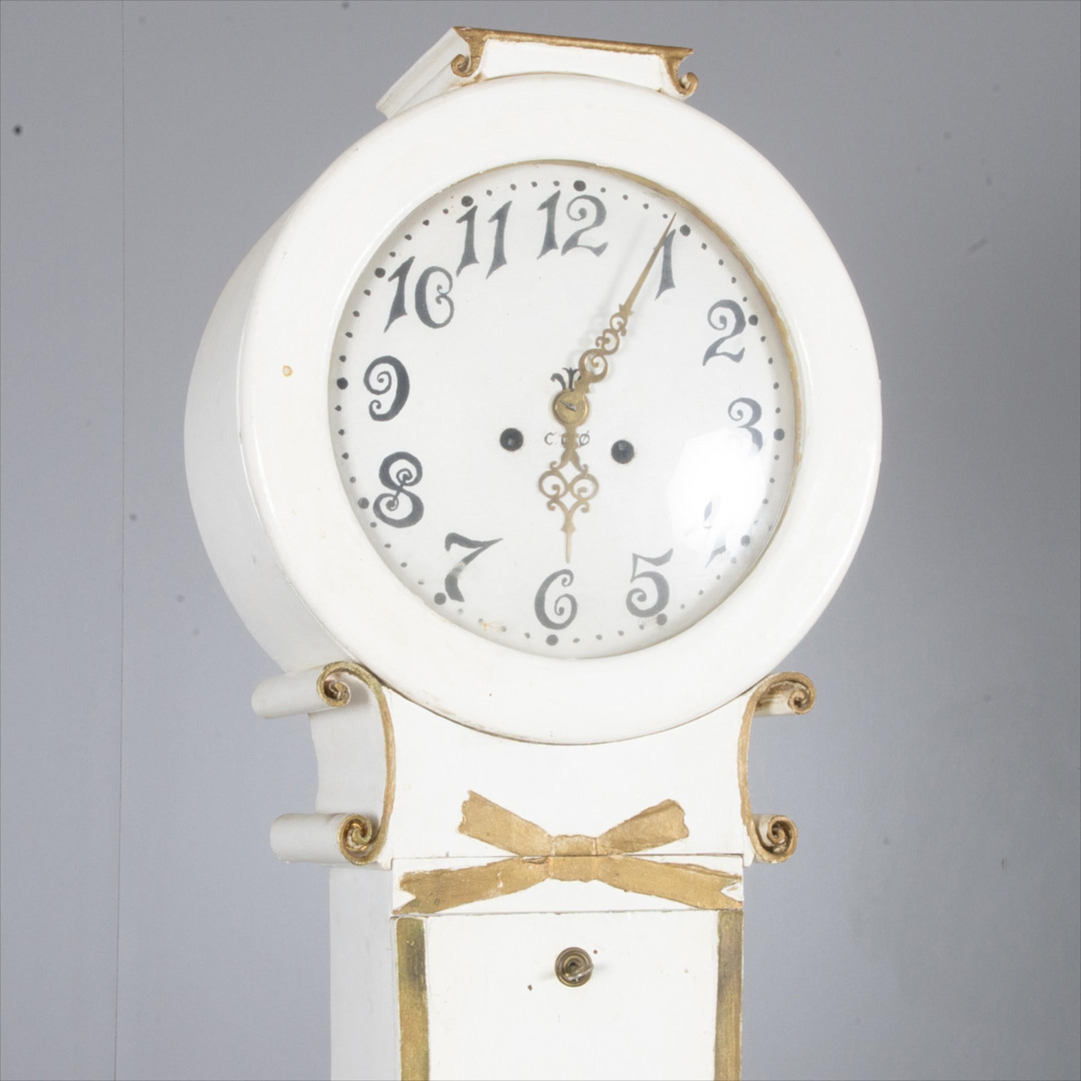 Swedish antique Mora clock in the Fryksdall school finished in White paint with gold edge detailing.

This unusual Mora clock has interesting scrollwork at the neck with a ribbon motif in the centre.

It stands 207cm tall approx and has a