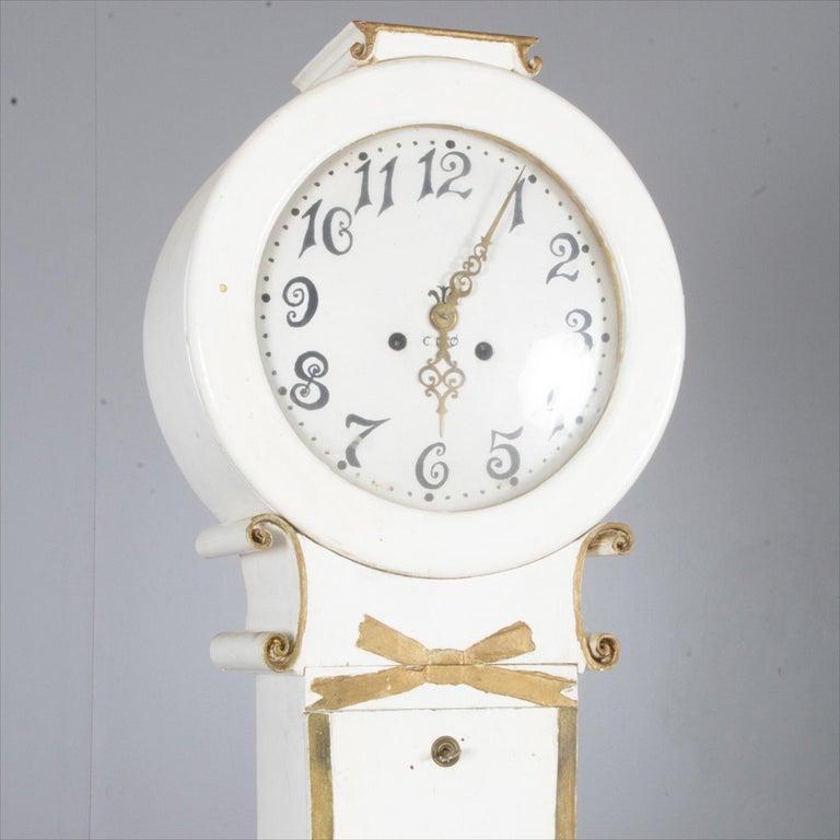 Swedish antique Mora clock in the Fryksdall school finished in White paint with gold edge detailing.

This unusual Mora clock has interesting scrollwork at the neck with a ribbon motif in the centre.

It stands 207cm tall approx and has a slender