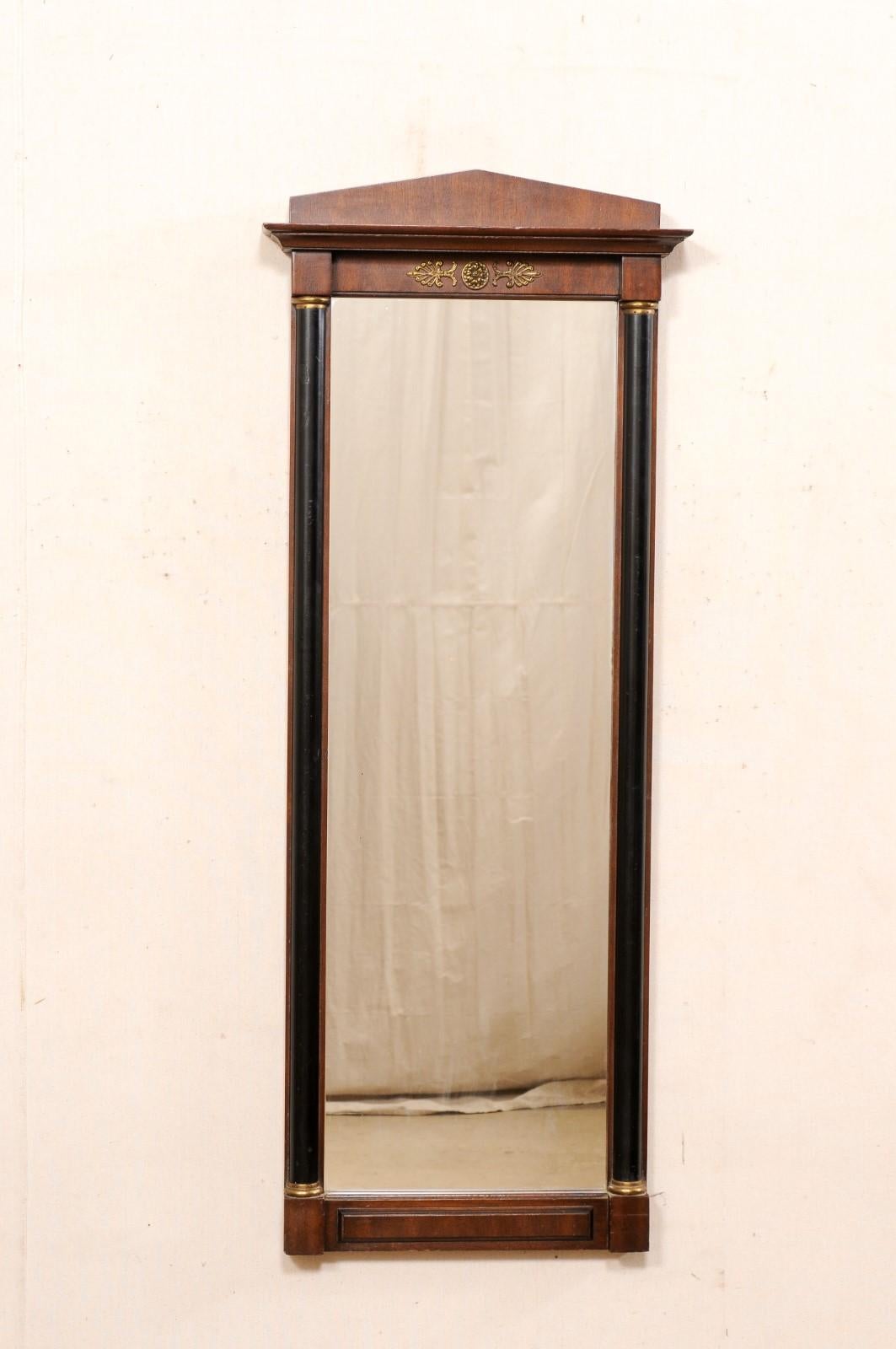 A Swedish Neoclassical-style wood mirror with brass accents from the early 20th century. This antique mirror from Sweden features a triangular pediment style crest with molded overhang adorning its top. The mirror is rectangular in shape, having a