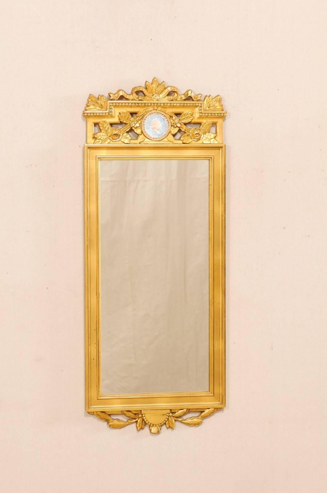 A Swedish neoclassical style gilt mirror from the early 20th century. This antique Swedish Neoclassical style gilded trumeau mirror features an ornately decorated pierce carved top in a bow and floral motif with beaded accents and a profiled cameo
