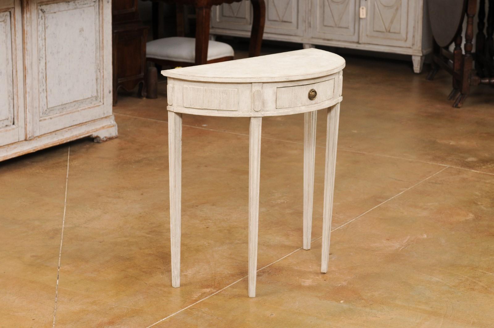A Swedish neoclassical style painted wood demilune console table from the 20th century with single drawer, carved fluted accents and tapered legs. Introduce an element of timeless elegance to your space with this Swedish neoclassical style demilune