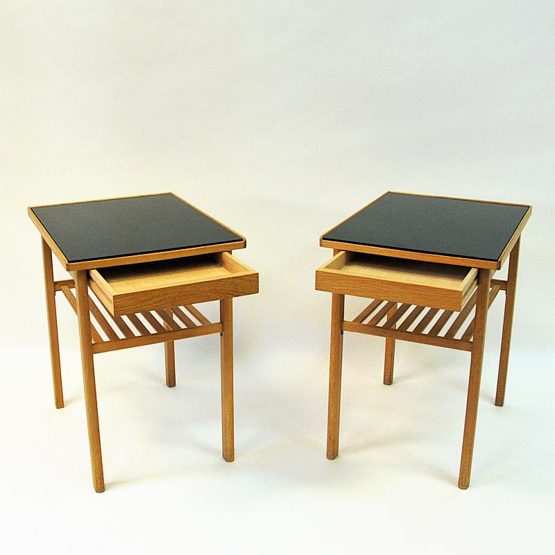 Lovely and Classic pair of vintage oak and glass top bed/ side tables from Svenska Möbel Industrier, 1960s, Sweden. Drawers underneath with a sprinkle shelf. These midcentury small tables has a black painted shiny glass plates on top. Good vintage