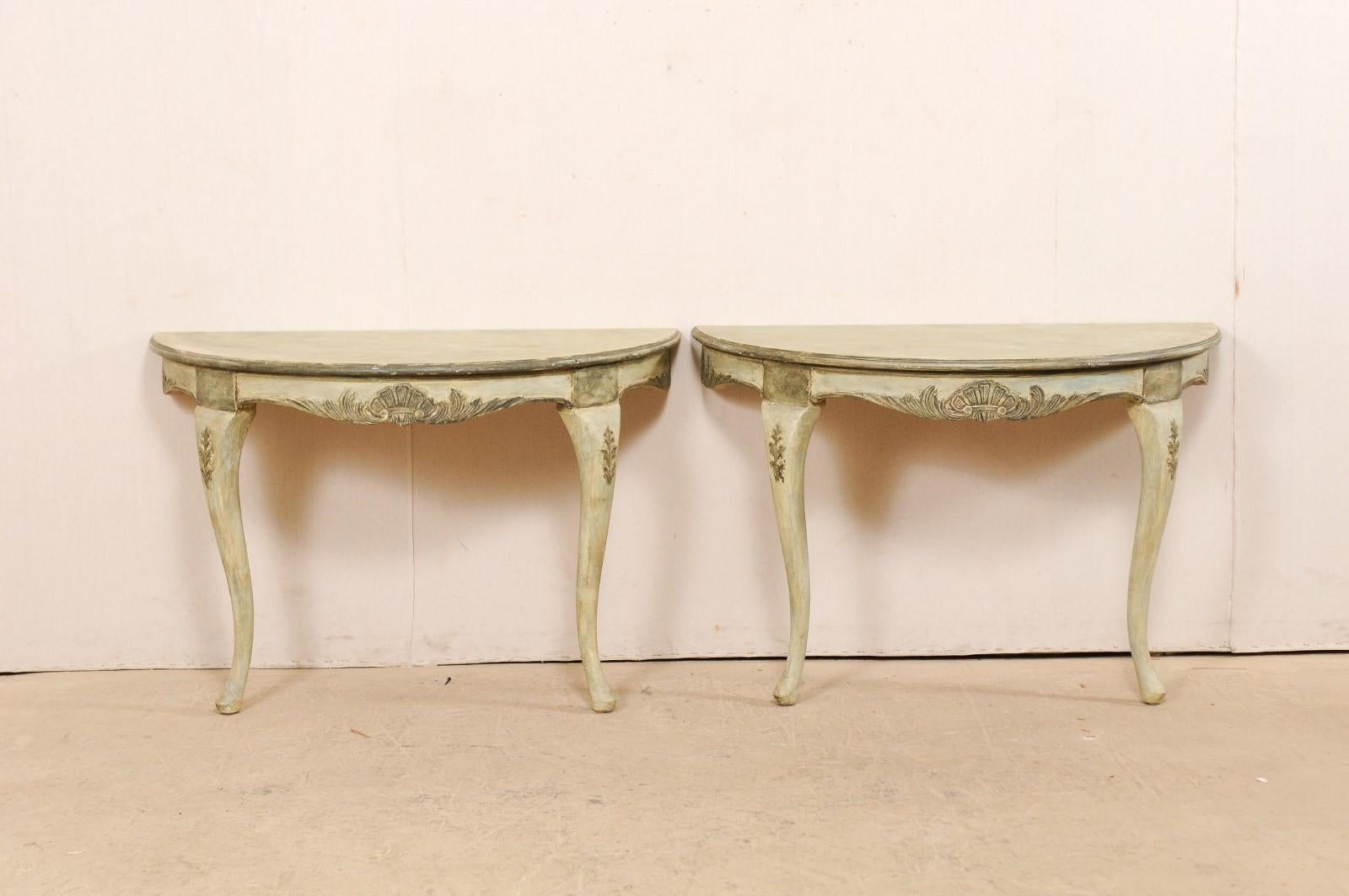 A Swedish pair of wall-mounted demilune tables from the mid-20th century. This vintage pair of wall consoles from Italy each feature a half moon top, over rounded aprons, with a carved decor of foliage motif and shell at front center. The tables are