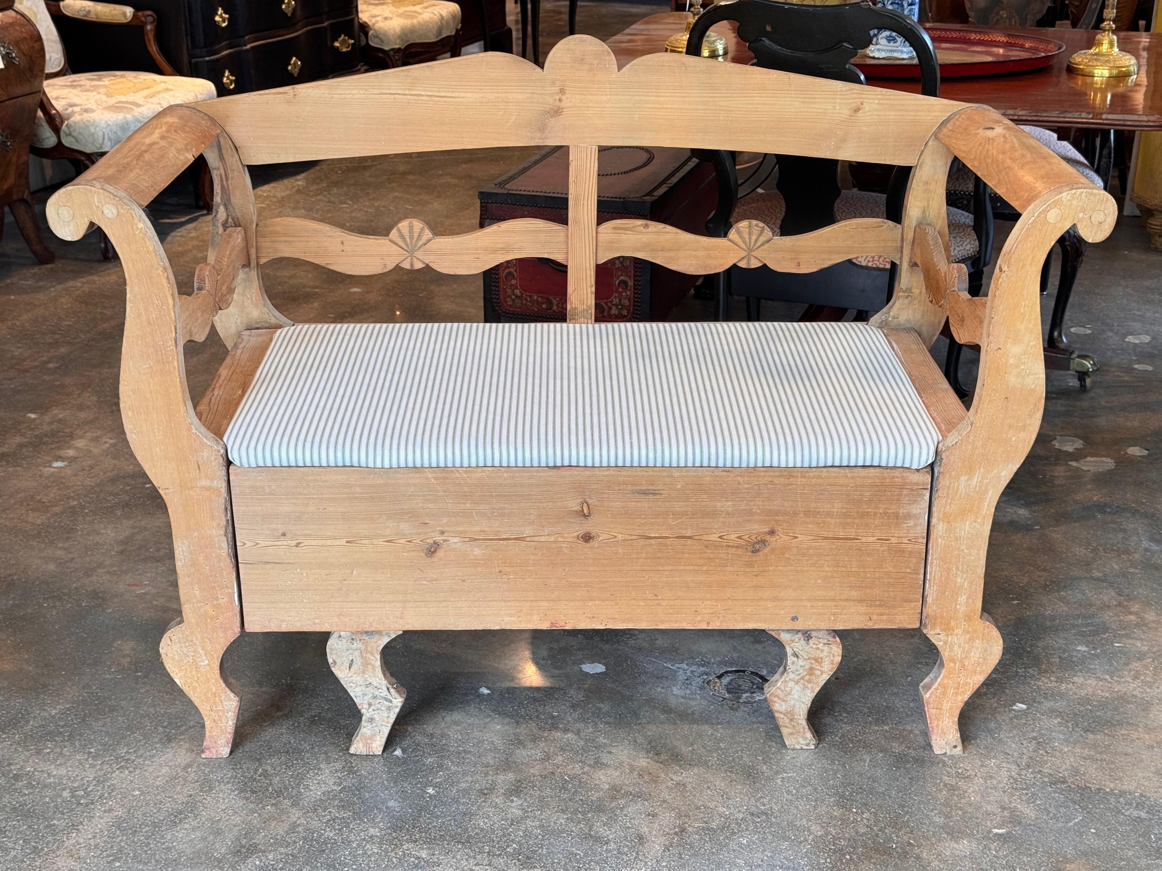 Rustic Swedish pine bench with a shaped backrest, curved arms, and splayed legs. The lift seat opens to reveal a generous storage compartment with visible dovetail joinery. Finished with a simple striped cushion, this piece combines practicality