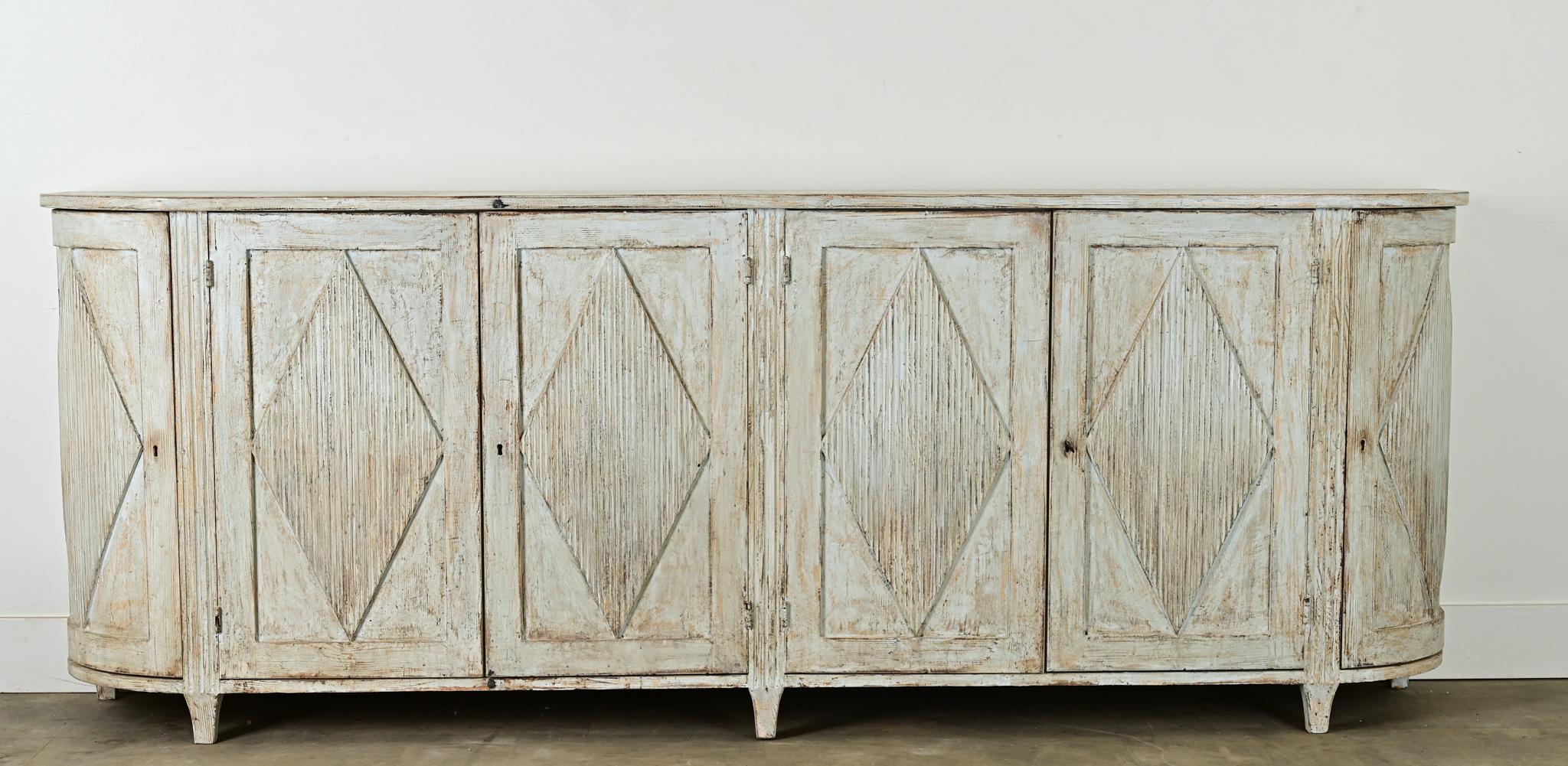A painted Swedish reproduction buffet has its original light gray-blue paint finish, worn in all the right places. The long solid pine cabinet has a shaped top over a row of paneled doors all with reeded diamond shape designs. The doors open with