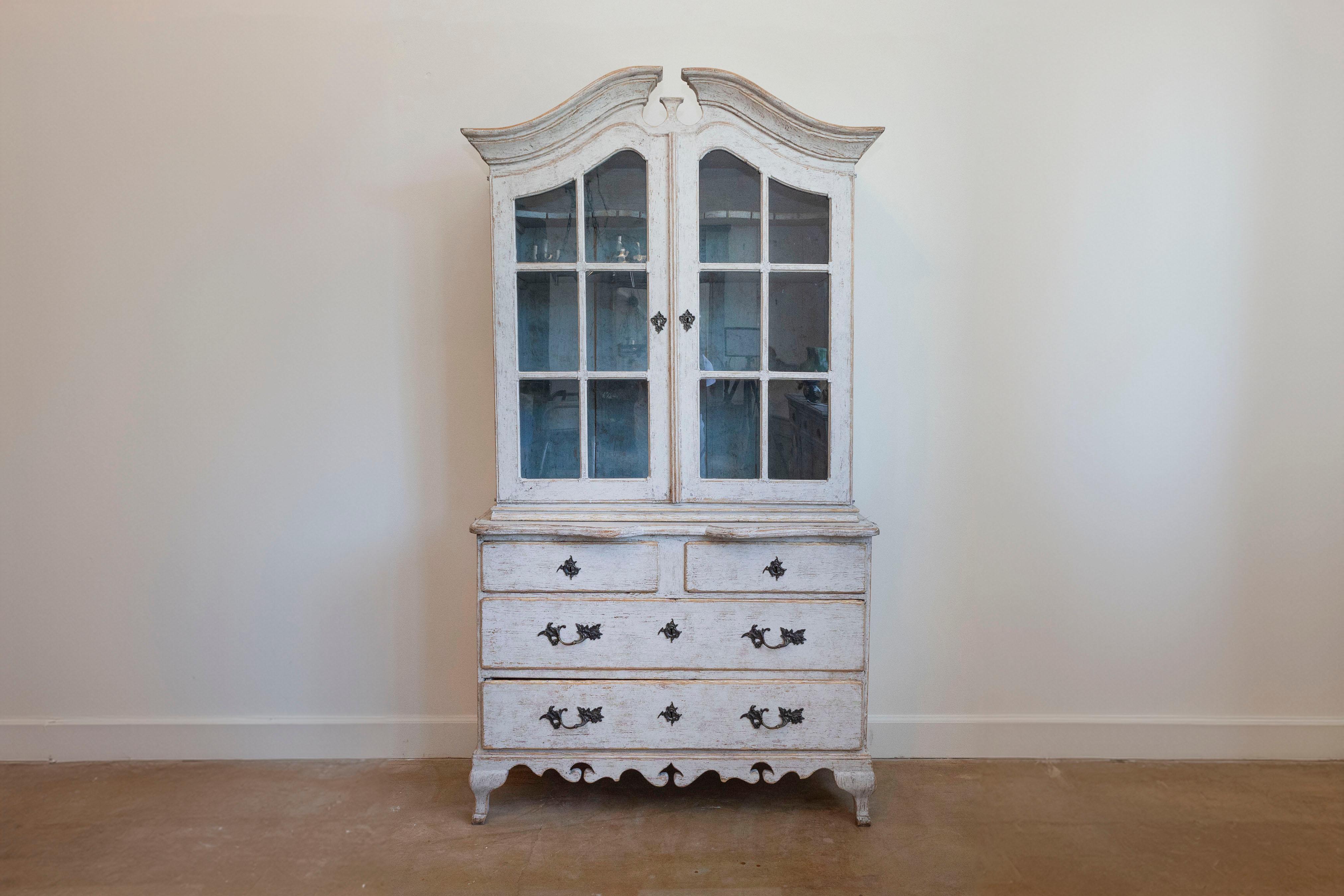 Mueble alto de estilo rococó sueco de hacia 1850 con tapa de bonete, acabado pintado de gris claro, puertas de cristal, cuatro cajones, faldón tallado y patas curvas. Sumerge tu hogar en la elegancia del estilo rococó sueco con este exquisito