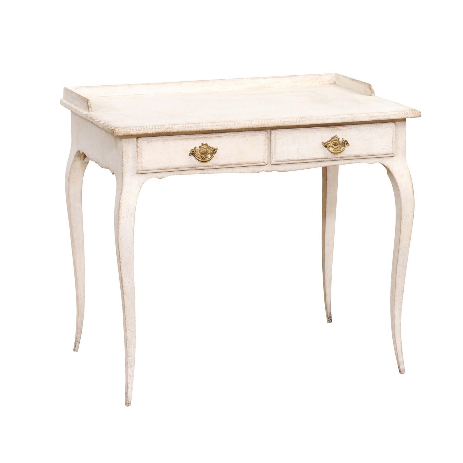 A Swedish Rococo style off white painted desk from circa 1880 with two drawers, cabriole legs and bronze hardware. This Swedish Rococo style desk, dating back to approximately 1880, exudes timeless elegance with its soft off-white/very light gray