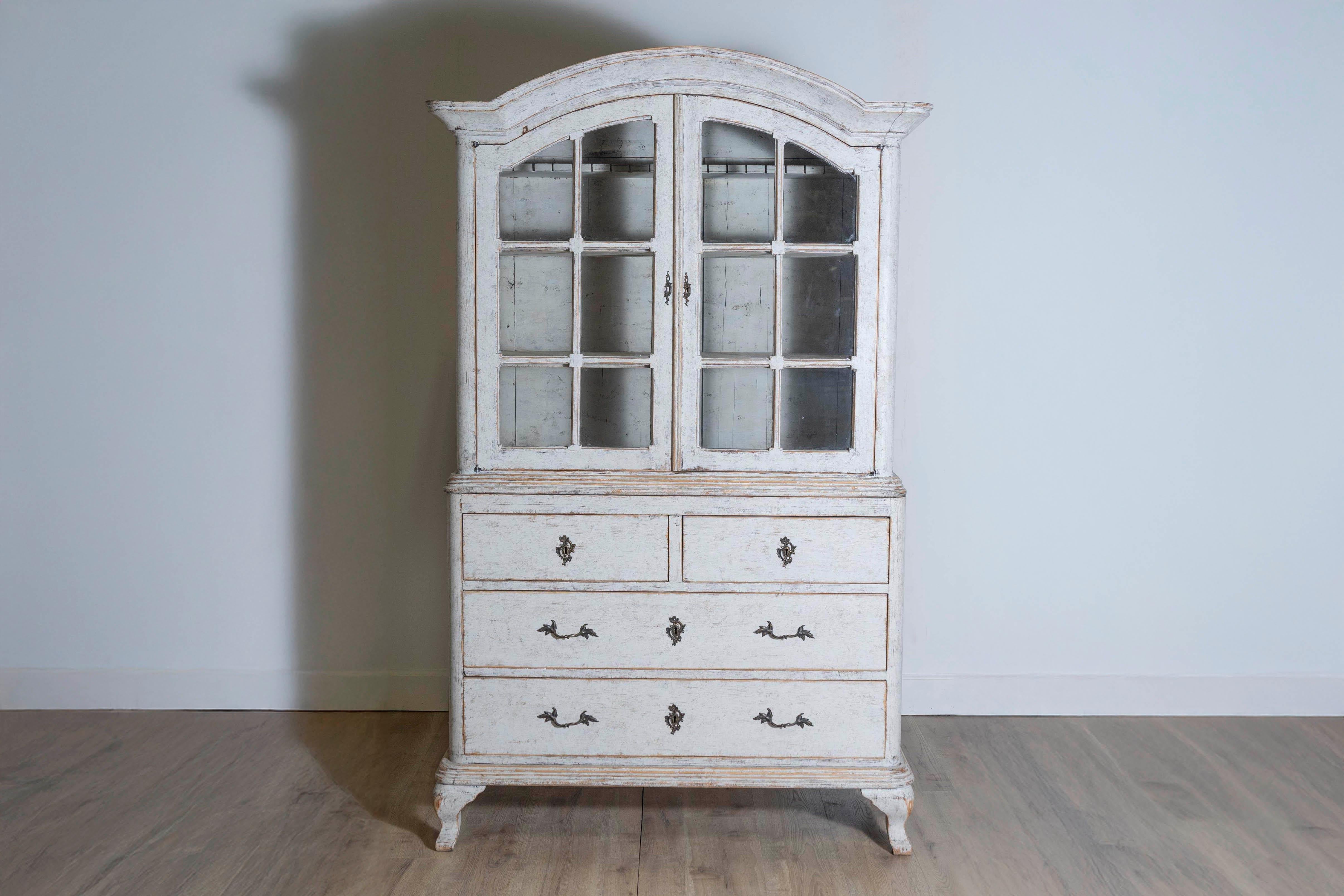 Bemalte Holzvitrine im schwedischen Rokoko-Stil aus dem 19. Jahrhundert, mit Haubenaufsatz und Glastüren. Diese exquisite Vitrine, die im 19. Jahrhundert in Schweden entstand, zeichnet sich durch ein haubenartiges Gesims aus, das über zwei Türen mit