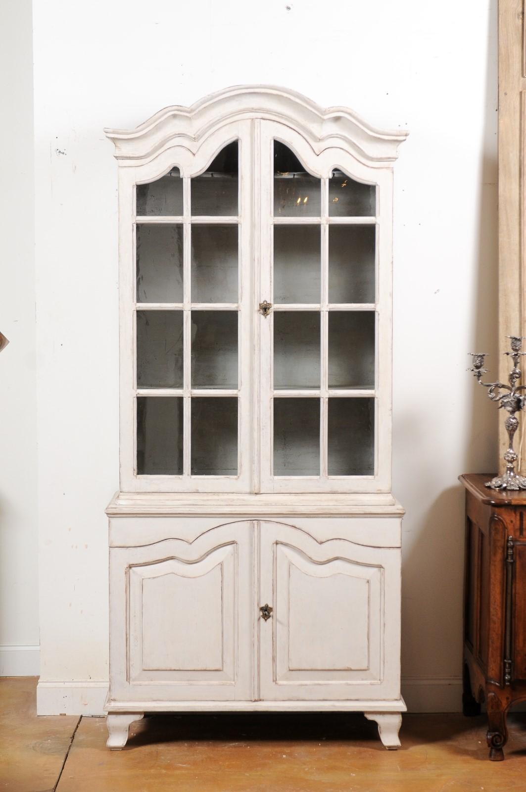 A Swedish Rococo style painted wood vitrine cabinet from the 19th century, with glass doors and arching accents. Created in Sweden during the 19th century, this cabinet features a molded arching cornice sitting above two paneled glass doors opening