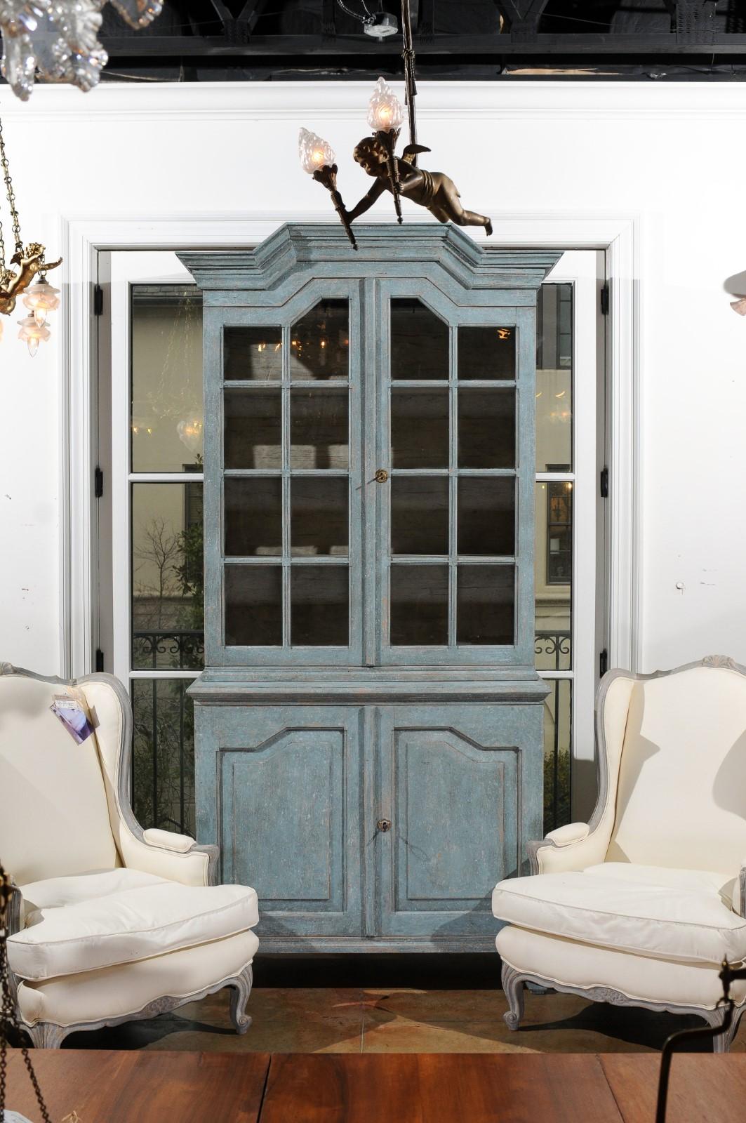 A Swedish Rococo style blue grey painted cabinet from the 19th century with linear pediment, glass doors and scrolling feet. Created in Sweden during the 19th century, this Rococo style cabinet captures our attention with its angular pediment