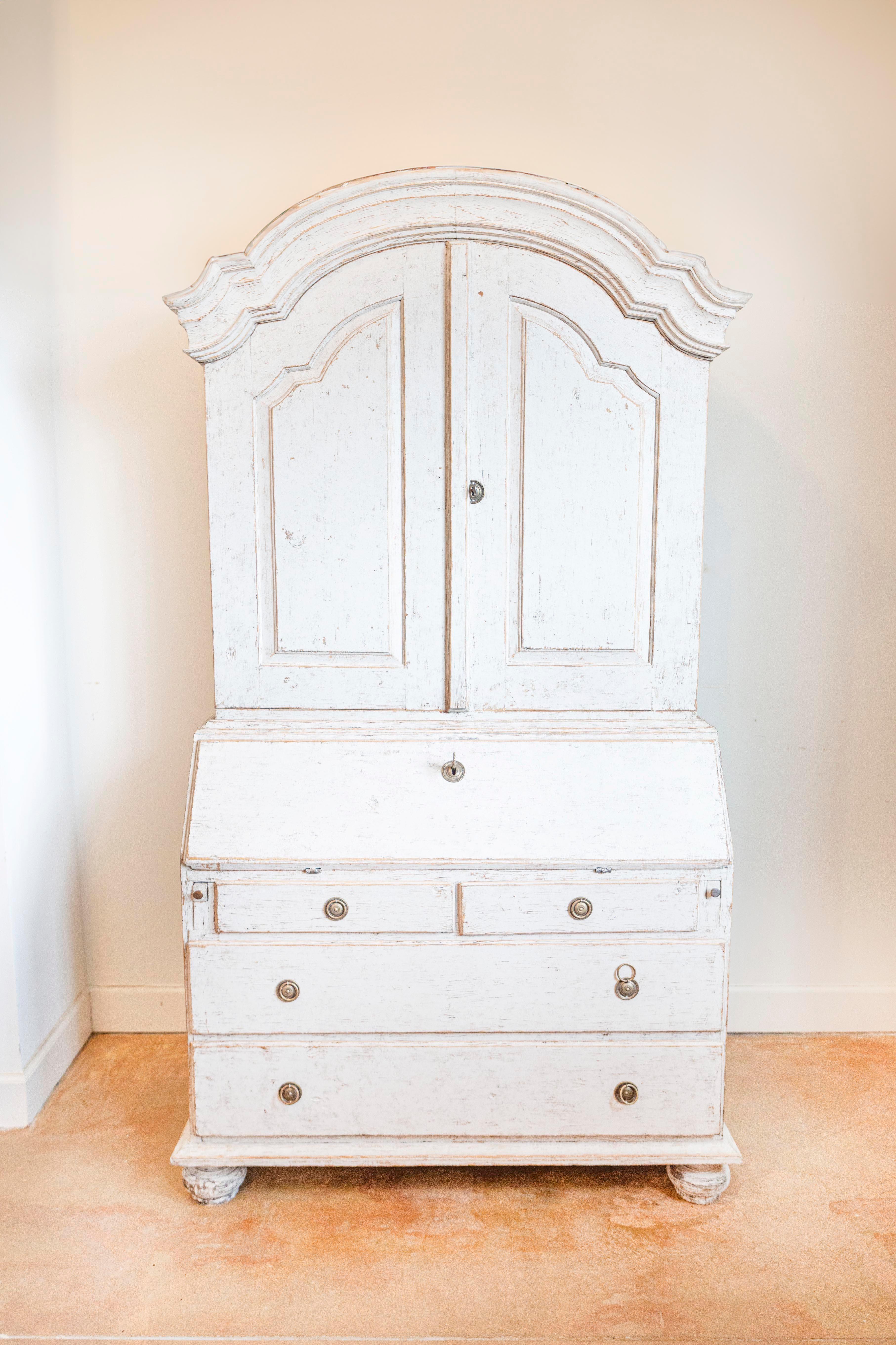A Swedish Rococo style tall secretary from circa 1820 with gray painted finish, chapeau de gendarme bonnet top, two doors, slant-front desk and four drawers below. This Swedish Rococo-style tall secretary, dating back to around 1820, is a testament