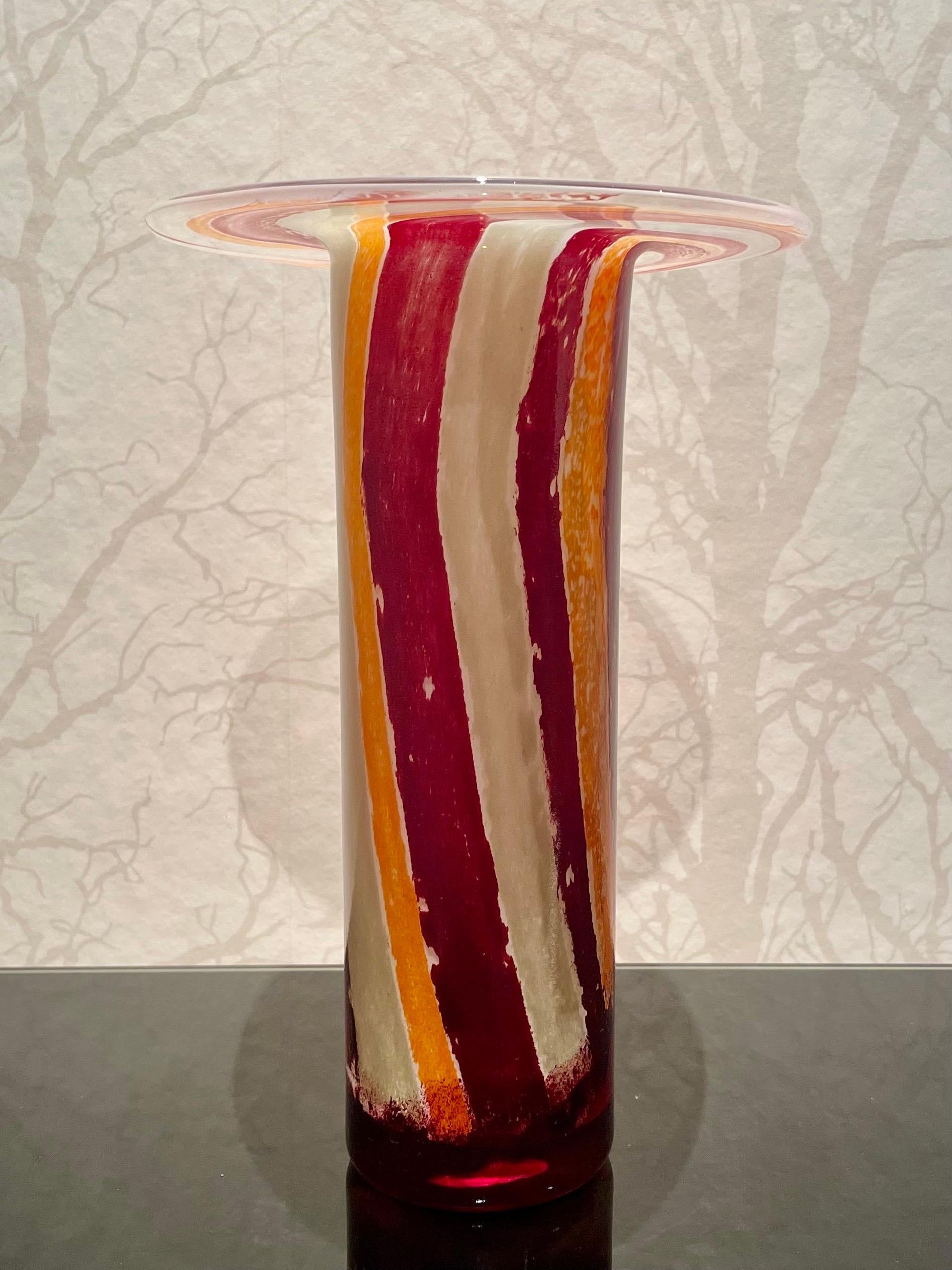 This is a colorful, sculptural glass vase by the Swedish glass artist Kjell Engman.
It comes in a tall cylinder shape.
It has a thick trunk and a thin, flat and very wide top like a brim of a hat. The pattern consists of vertical, slightly twisted