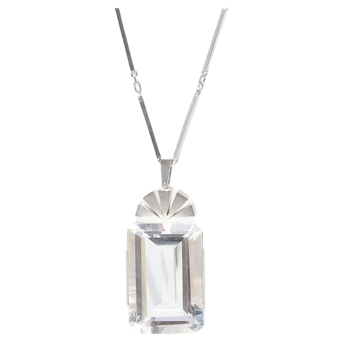 Questa collana in argento e cristallo di rocca presenta le caratteristiche tipiche di questo periodo, come la catena ad anelli e barrette e il ciondolo dalle forme geometriche rettilinee, la superficie lucida e il cristallo di rocca tagliato a