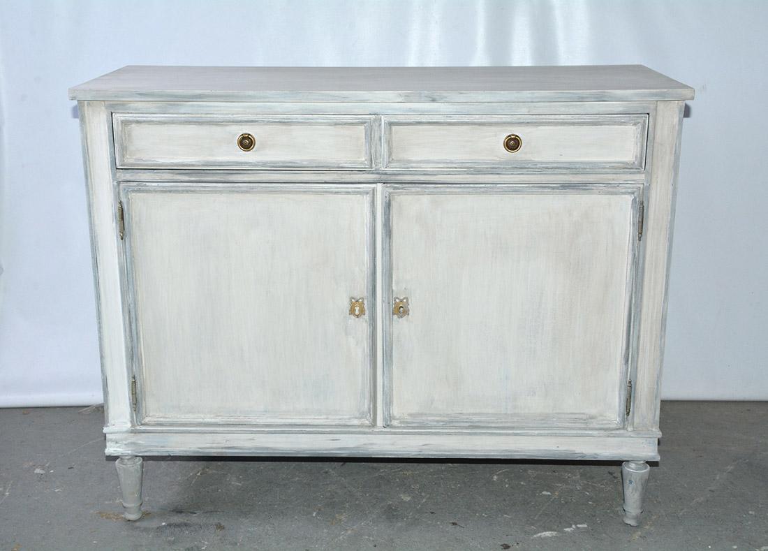 The Swedish style painted wood storage cabinet features a simple design of clean lines and has a lovely blue grey paint and subtle accented trim. There are two top drawers with Classic brass pulls over two large doors. The doors open to reveal