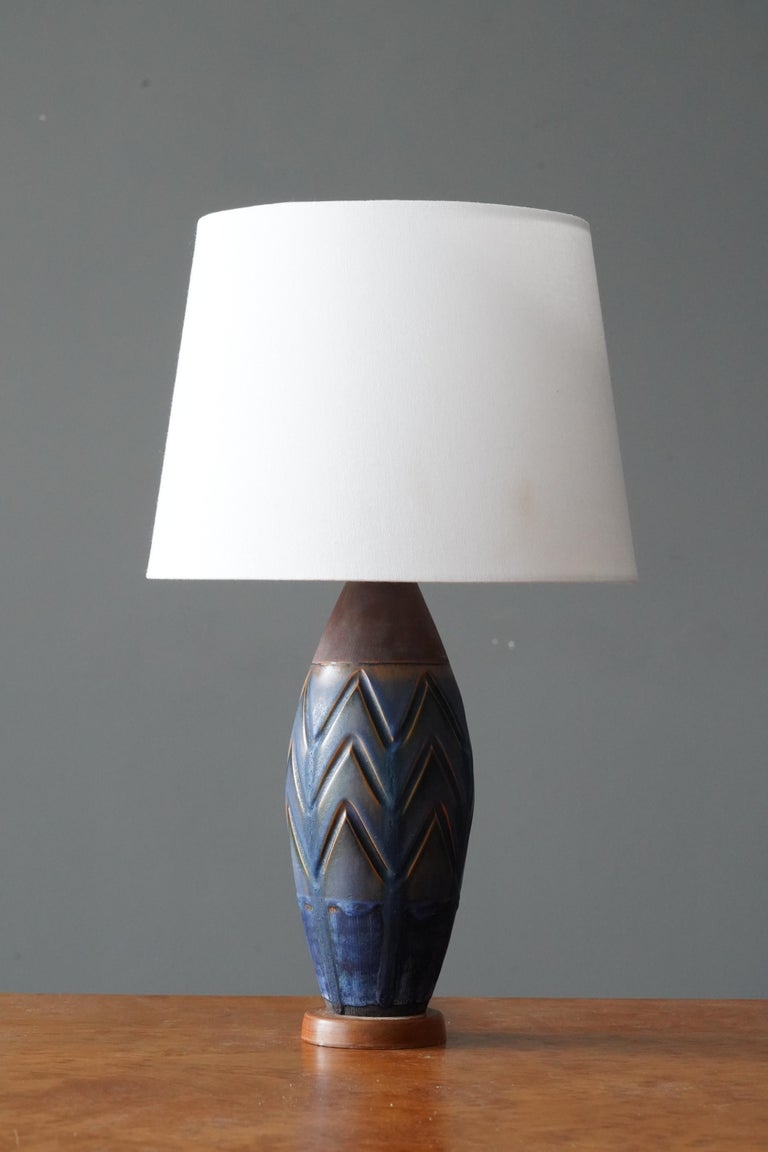 Table lamp, in incised stoneware, on wooden base. Glaze features brown and blue paint.

Dimensions listed are without shade. 
Dimensions with shade: height is 16.75 inches, width is 10 inches.
Dimensions of shade: top diameter is 7.5 inches,
