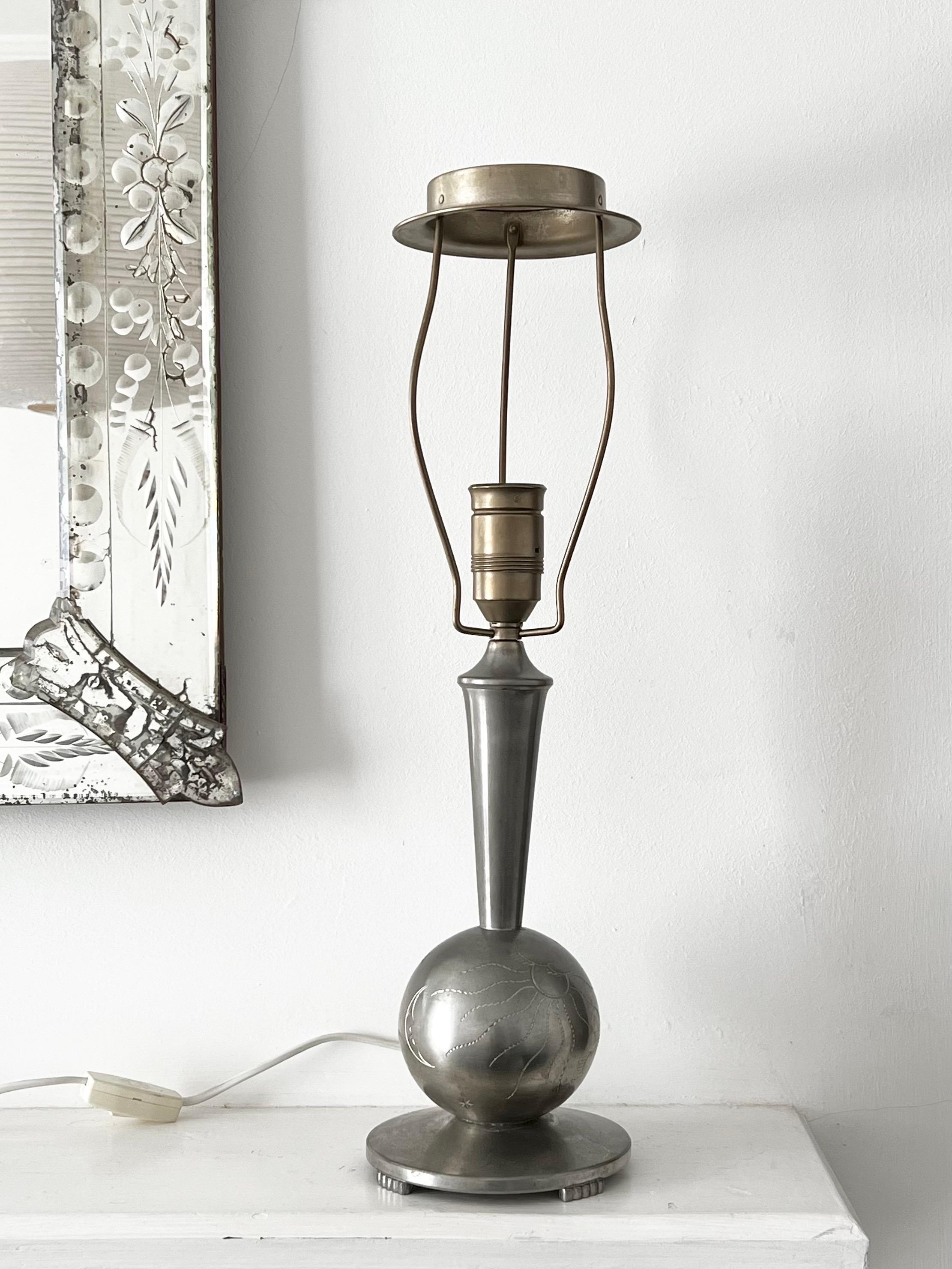 A pretty table lamp of white metal (pewter or tin) made in Sweden, 1930s. Swedish Grace, or Art Deco style. 

The design features a circular base raised on four feet, and central ball with engravings depicting the sun, moon, Saturn and lightning.