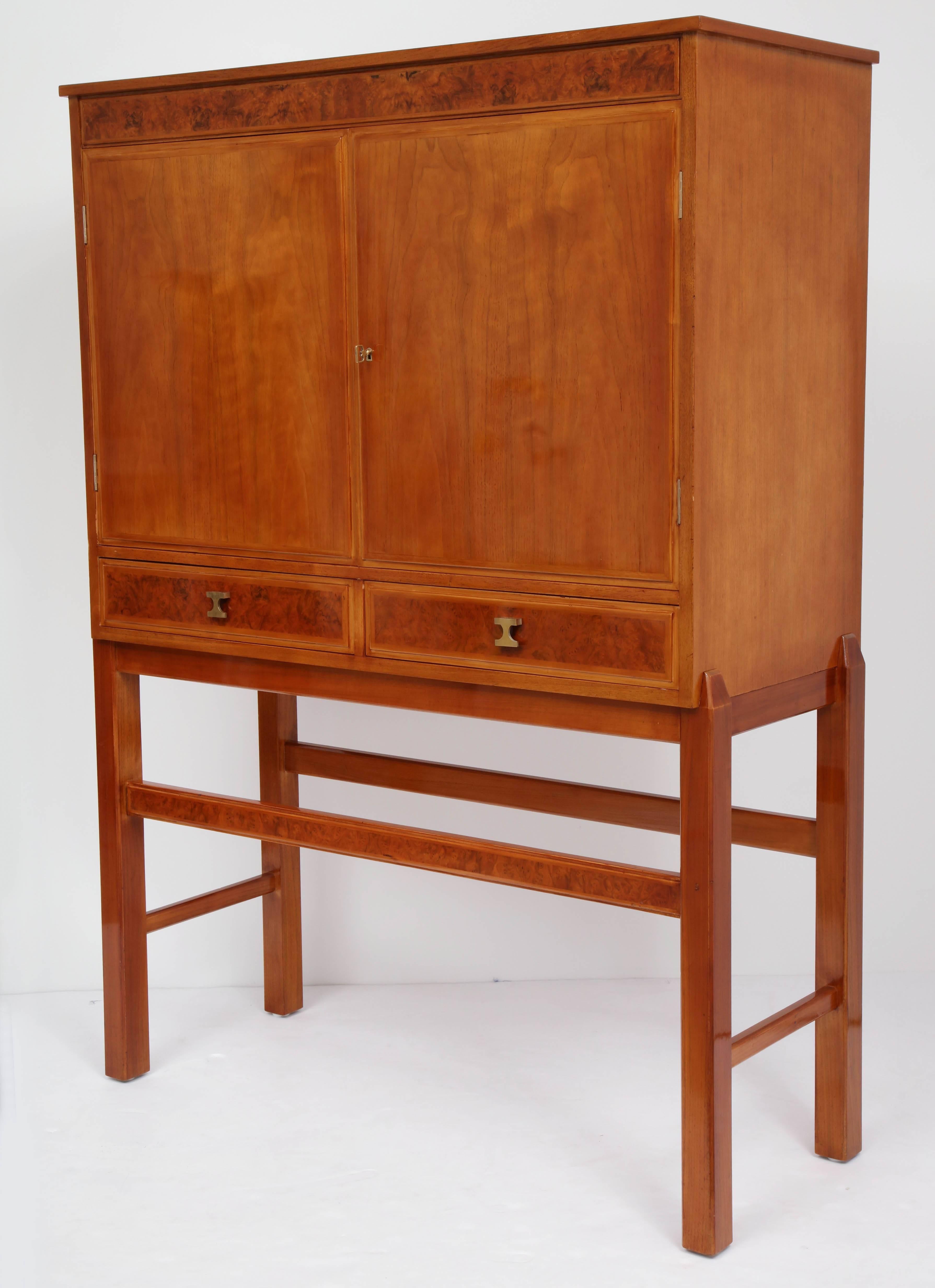 A Swedish walnut and burl walnut dry bar, circa 1940s, the rectangular body with two raised panel cupboard doors opening to a mirrored interior with a single half depth shelf, the doors with glass holders. Two lower short drawers, raised on a square