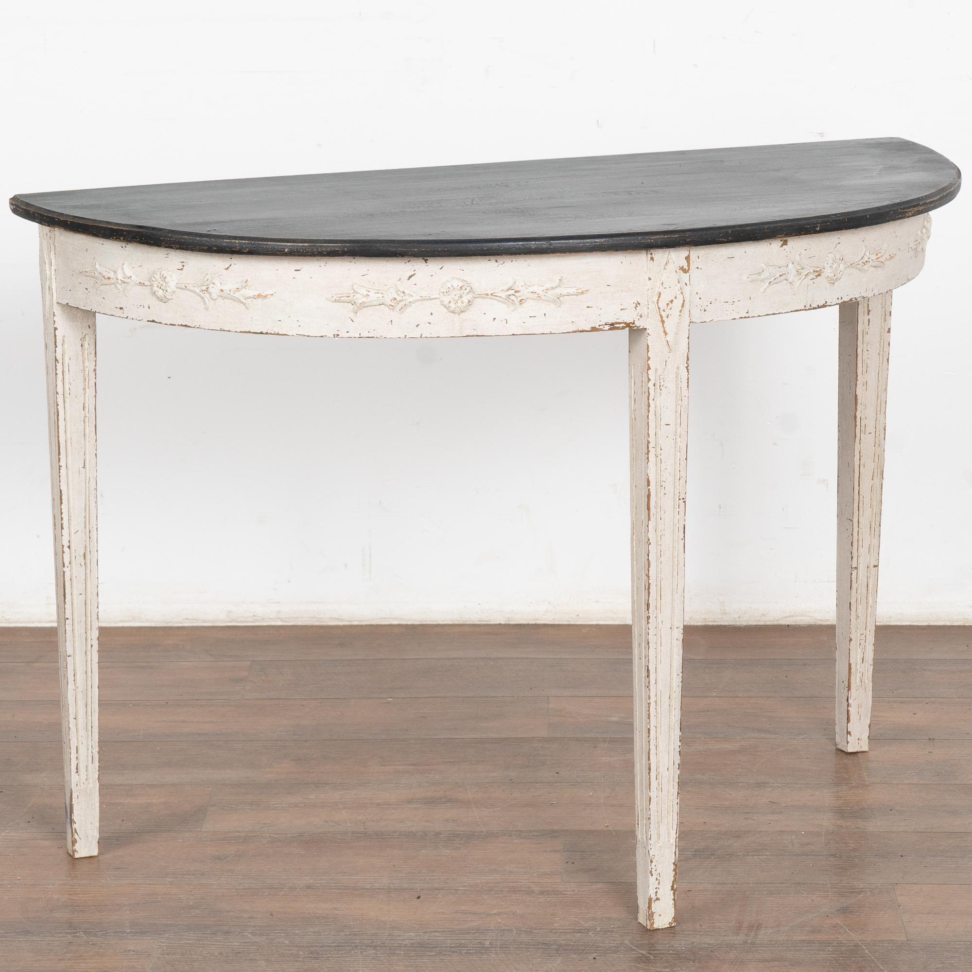 Gustavian demi-lune pine side table or console with gently tapered fluted legs and decorative applied carving along skirt.
Newer professionally applied layered antique white painted finish with contrasting black top, slightly distressed to fit age