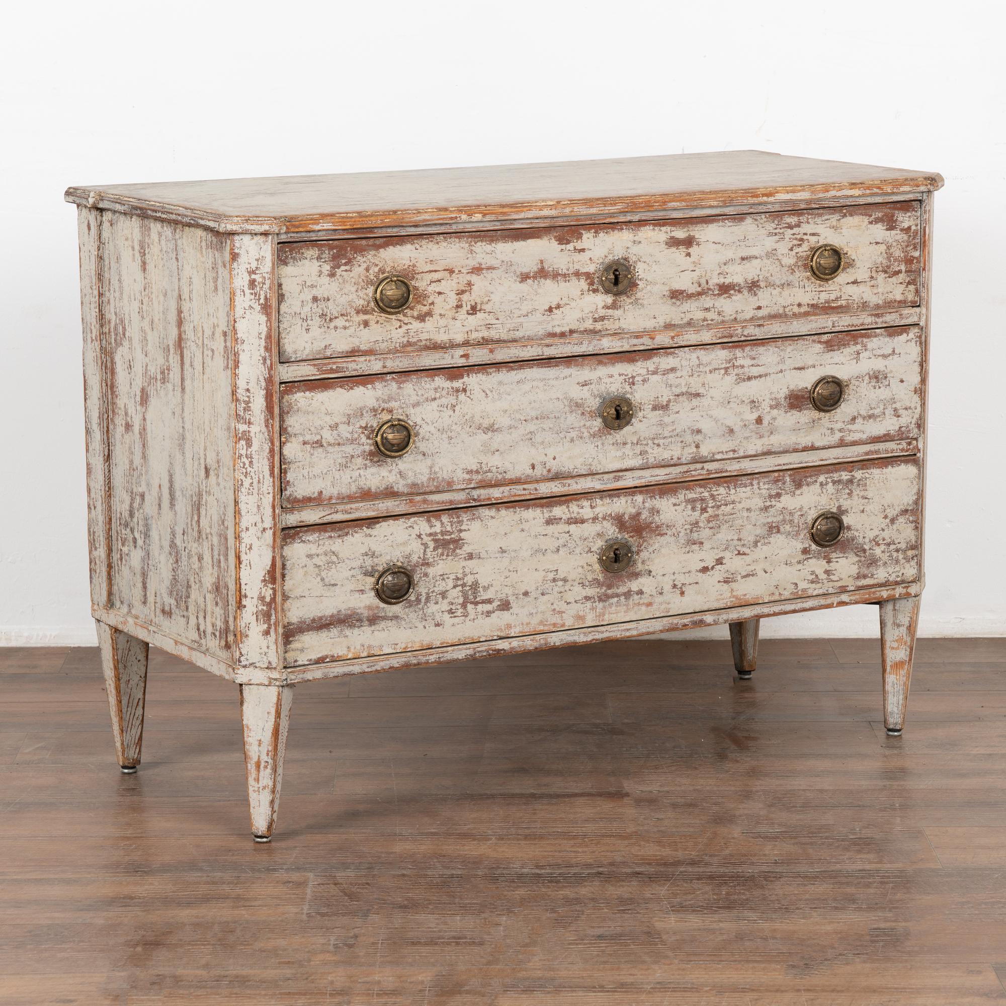 The simple lines of this charming pine chest of three drawers with canted sides and tapered feet are reflective of its Swedish country styling. 
The newer, professionally applied layered antique white painted finish with brick red and gray