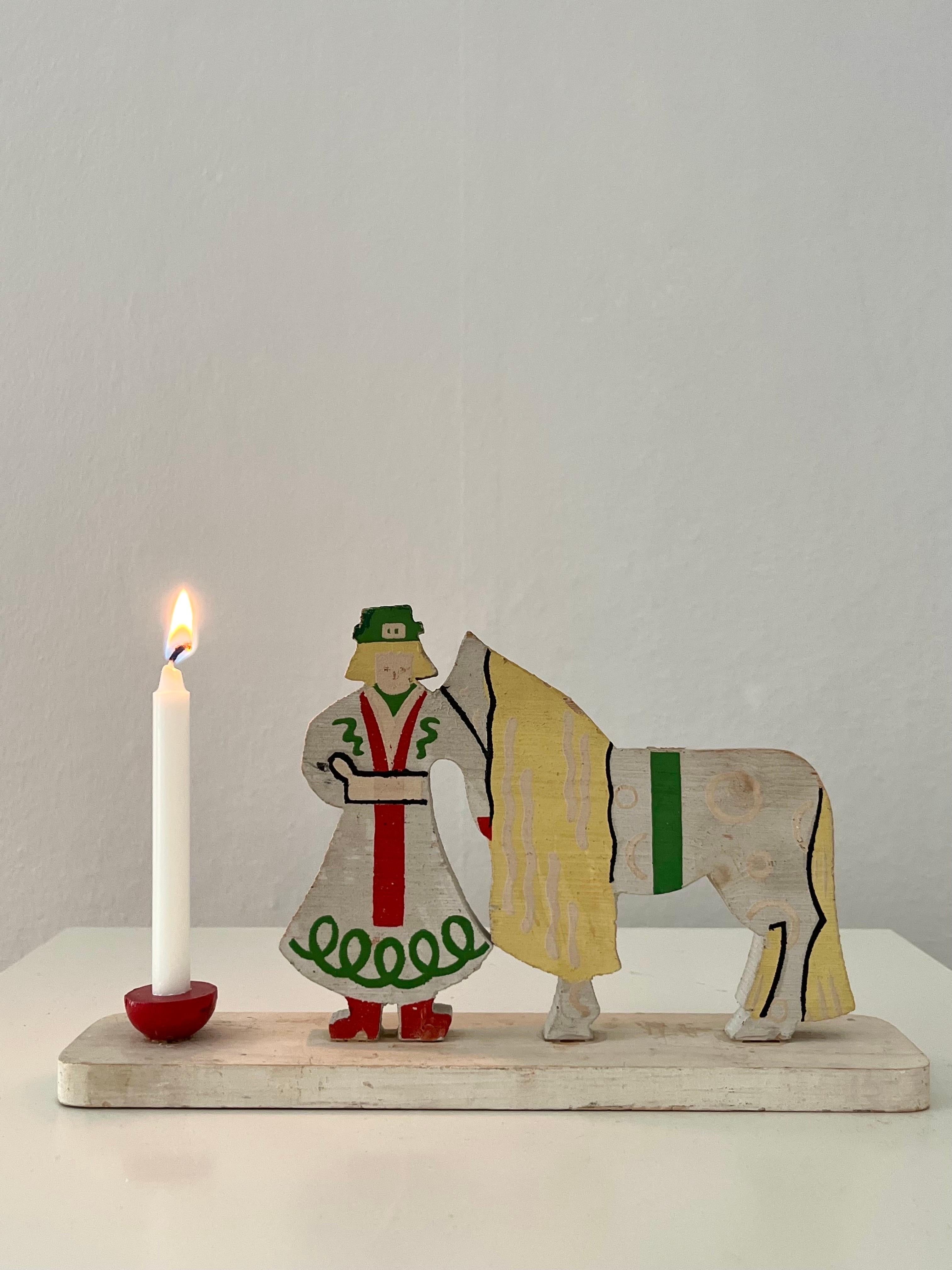 Understated, yet significant Christmas candle holder from Sweden in the 1950s/1960s. On a wood base is a wooden carving depicting a woman and a horse painted in green, red, yellow and black on a white base. In on end is a red candle holder. 
